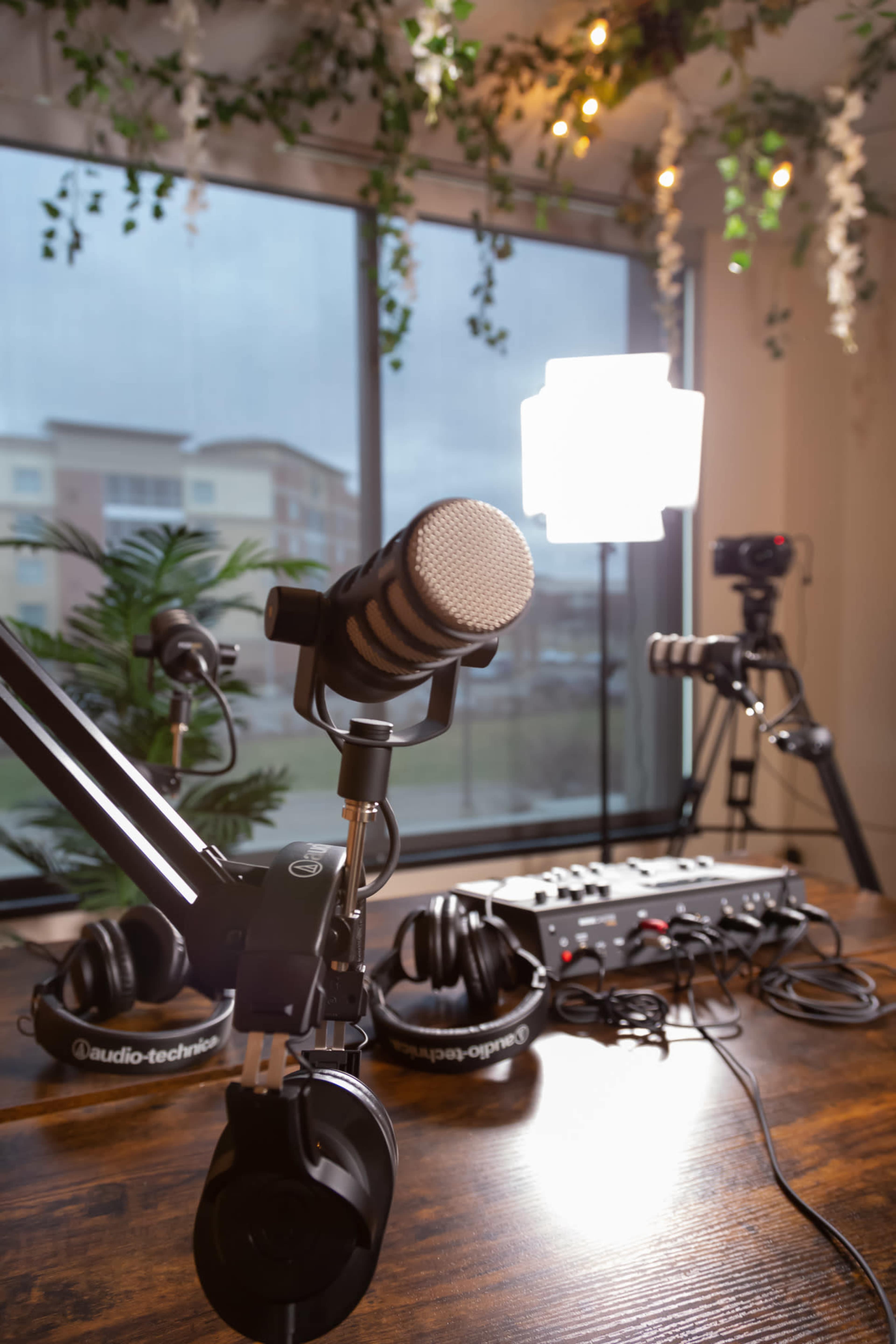 A podcasting setup with a microphone, headphones, and lighting equipment arranged on a wooden table near a large window.