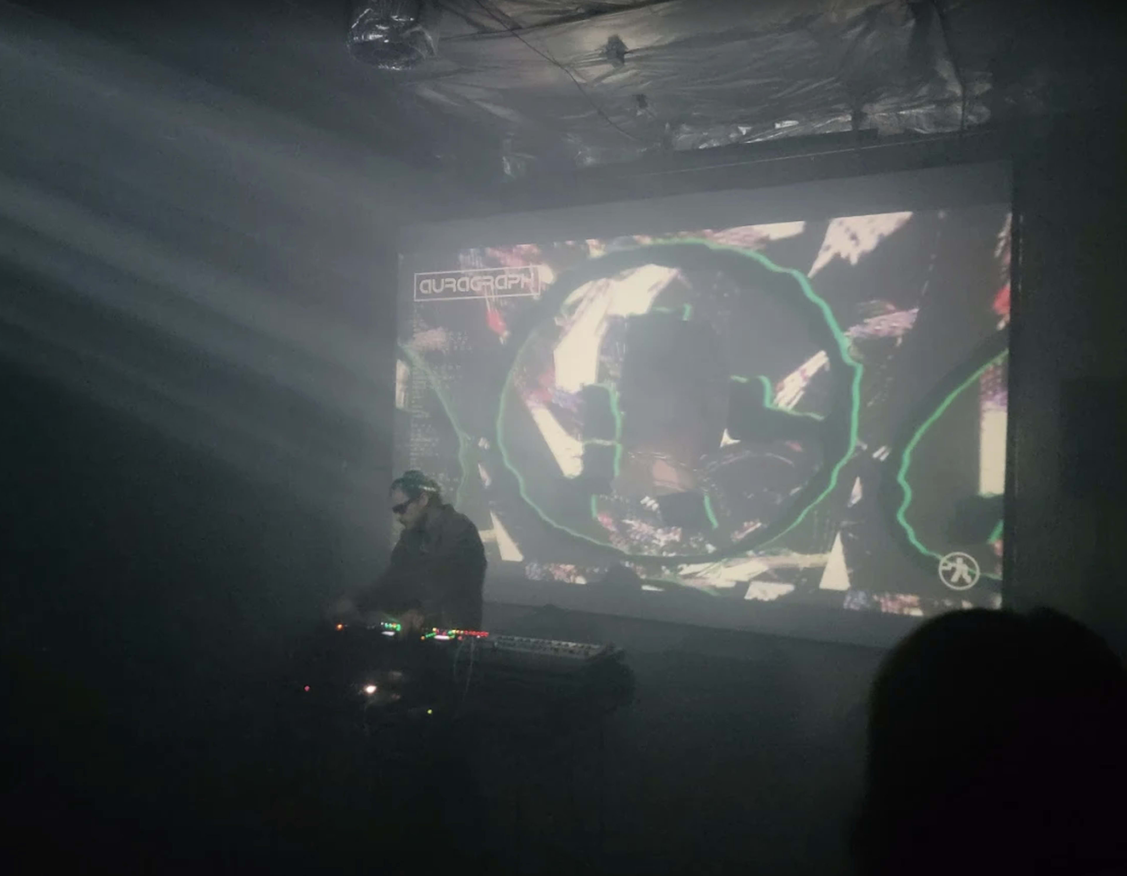 A musician performs in a dark venue with a large projection displaying abstract visuals in the background.