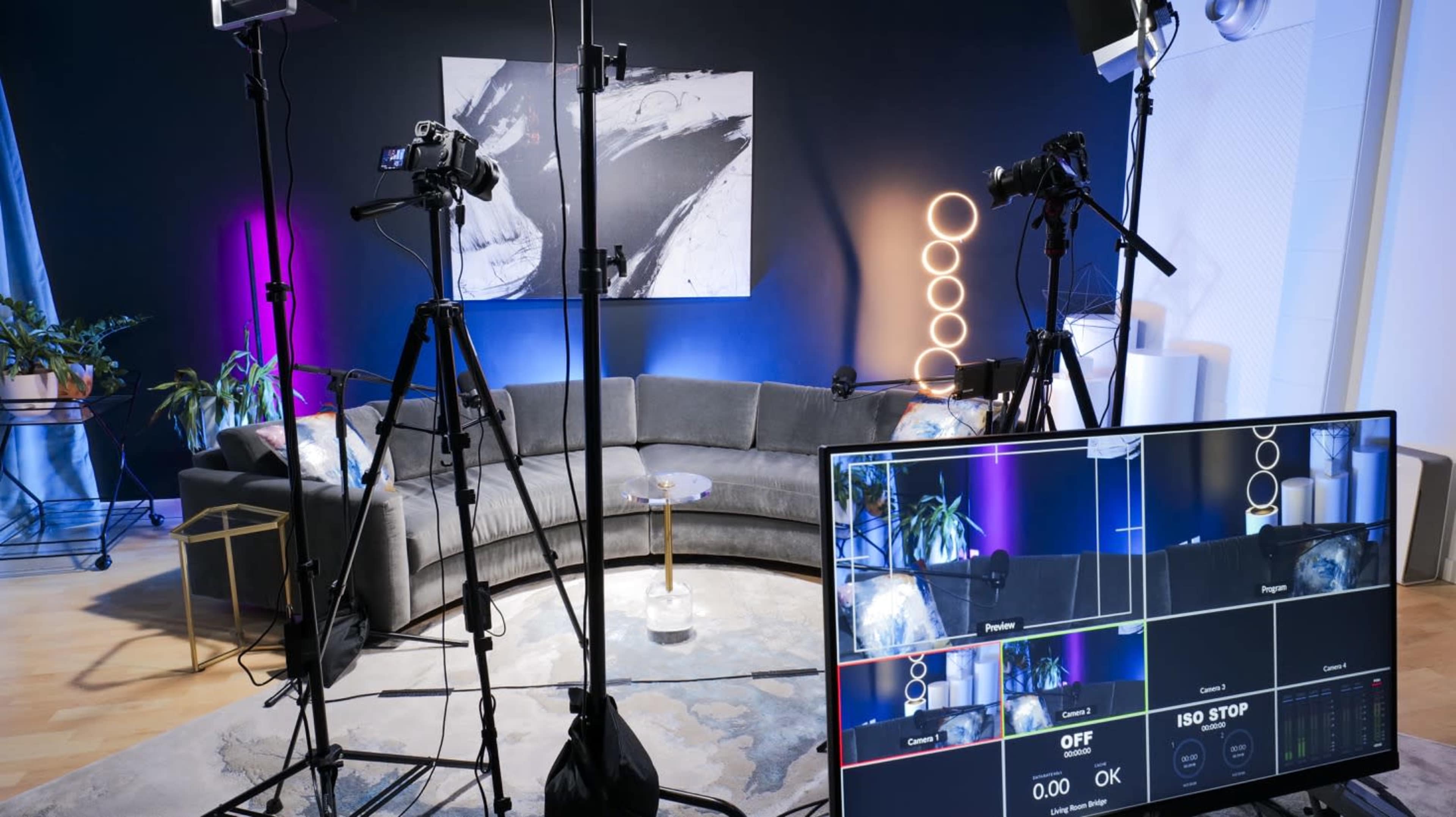 The image shows a modern studio setup featuring a curved couch, multiple cameras on tripods, and a large monitor displaying a broadcasting interface.
