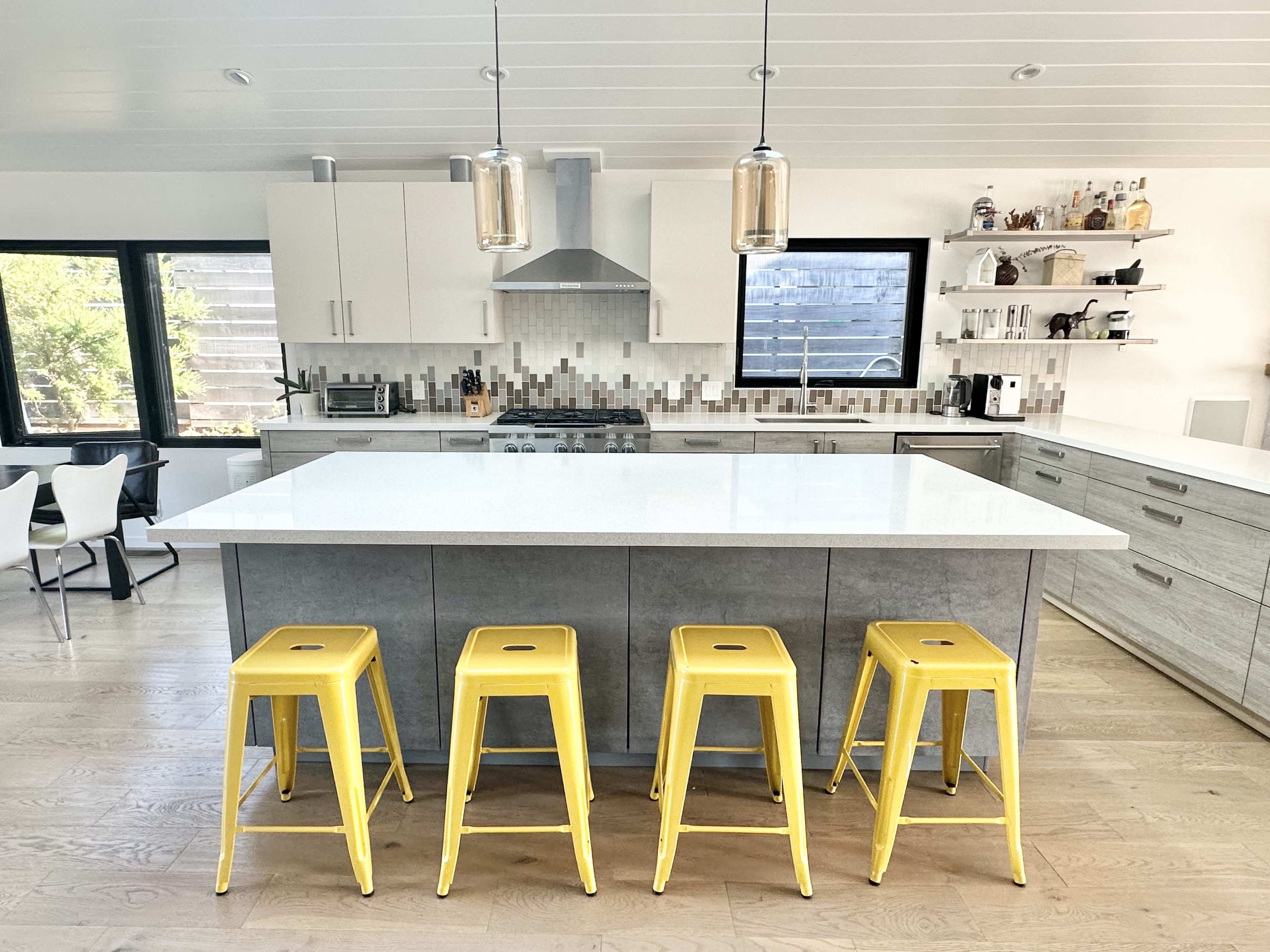 A modern kitchen with a centered gray island, four yellow stools, and a minimalist design featuring white cabinets and a backsplash of neutral tiles.