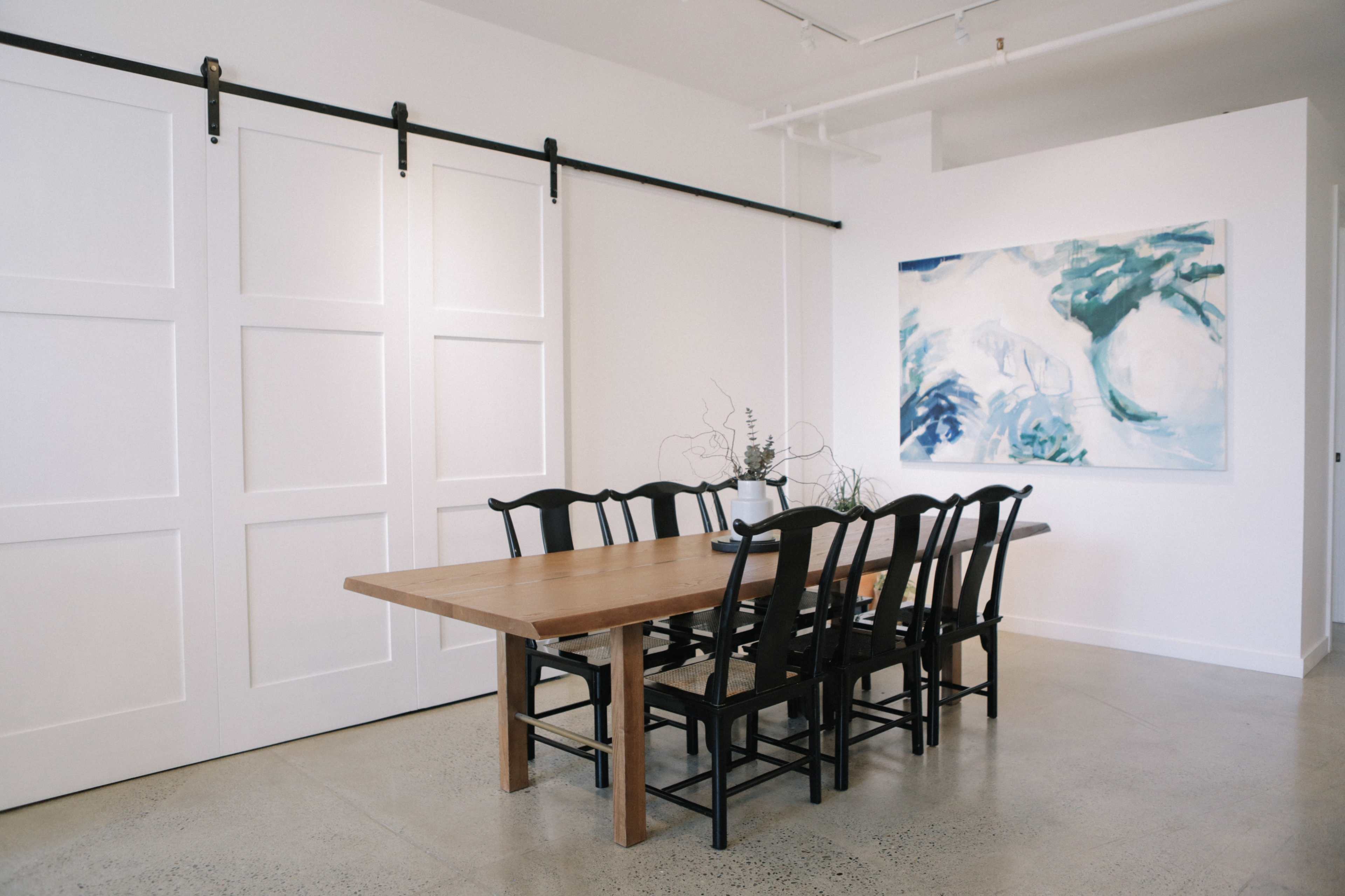 The image shows a minimalist dining area with a wooden table surrounded by black chairs, a large abstract painting on the wall, and sliding barn doors in the background.