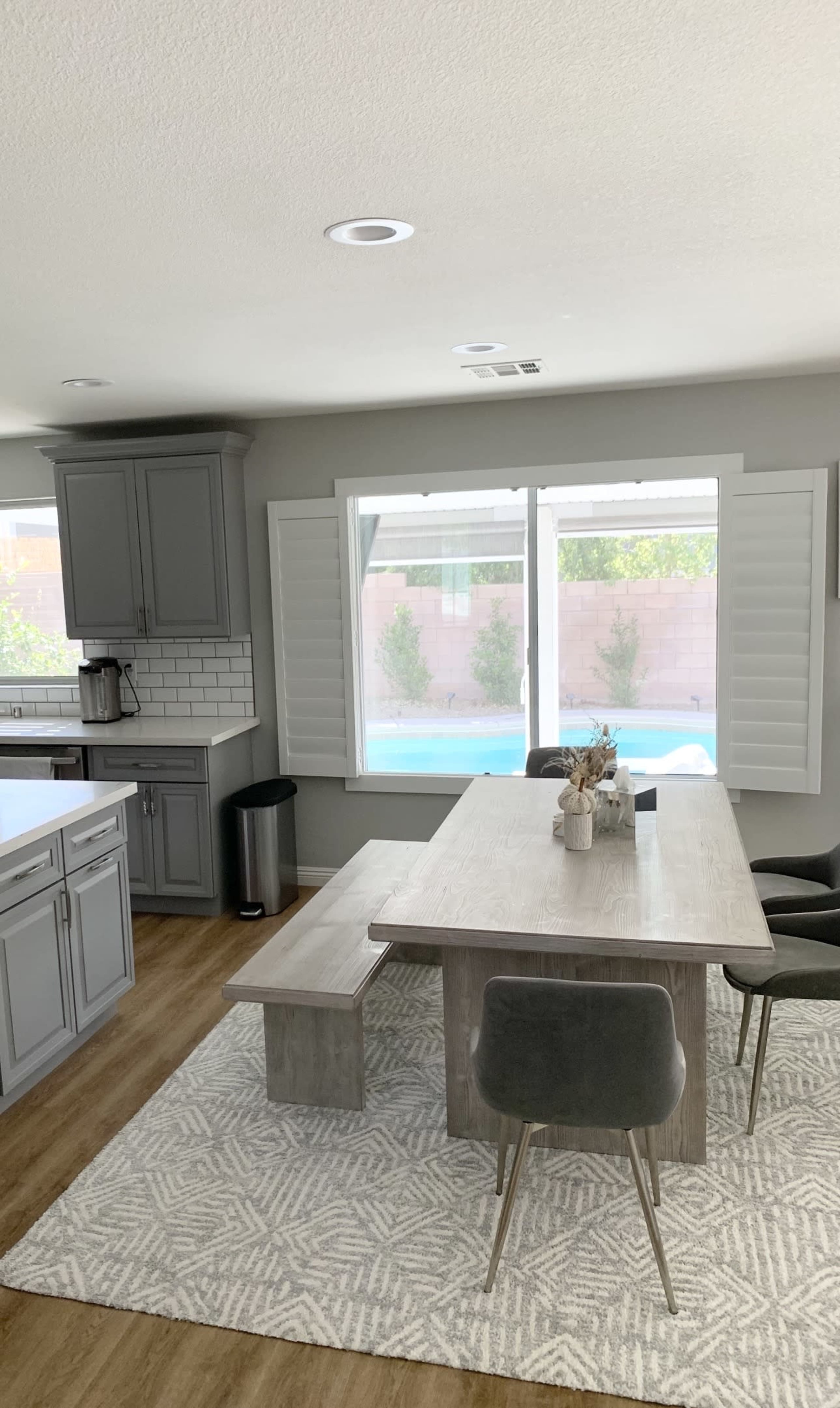 A dining area with a rectangular table, benches, and chairs is situated next to large windows overlooking a pool, with gray cabinets in the kitchen area.