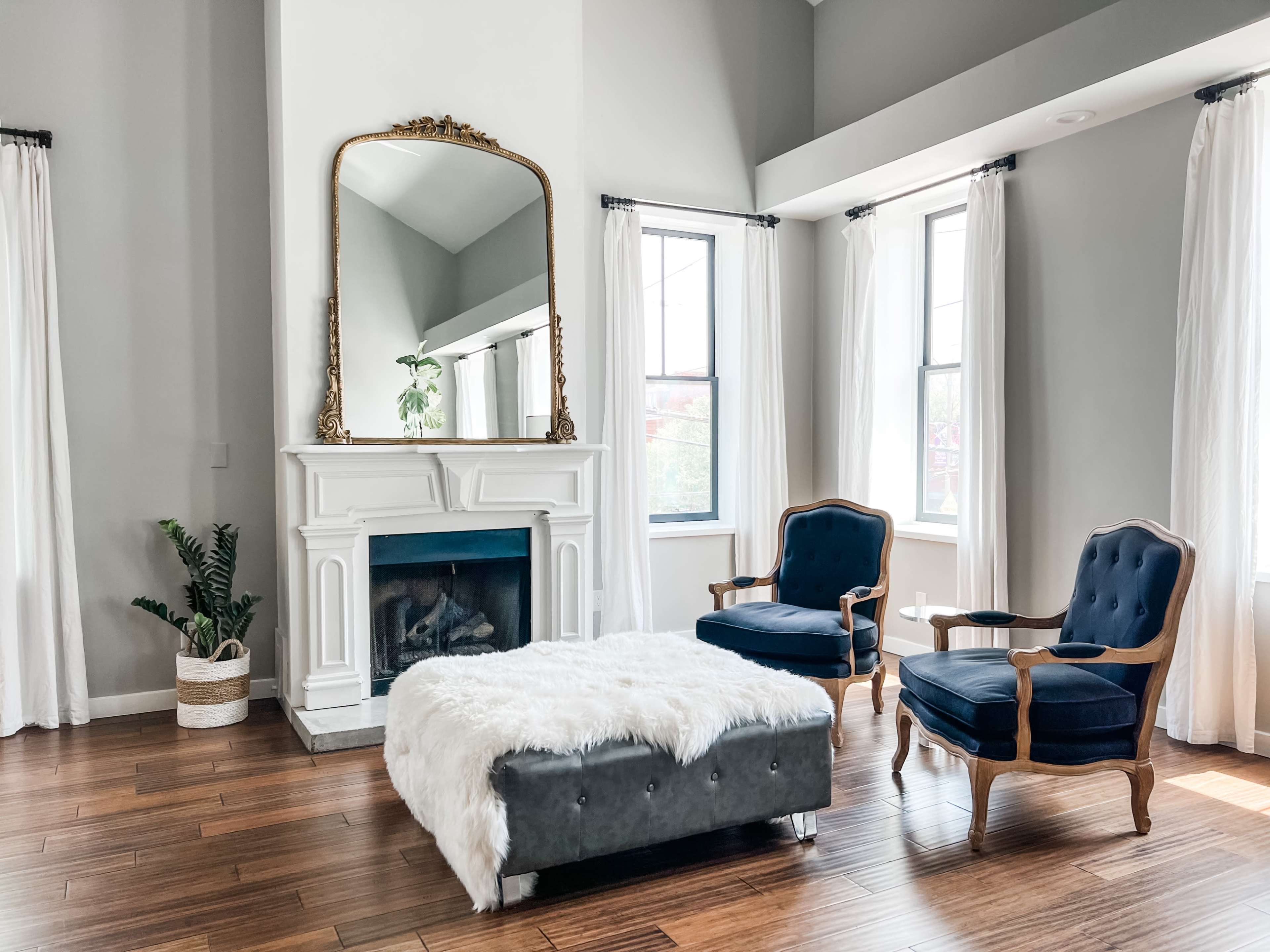 A living room with a white fireplace, a large mirror, two blue armchairs, and a fur-covered ottoman, all set against light gray walls and large windows.