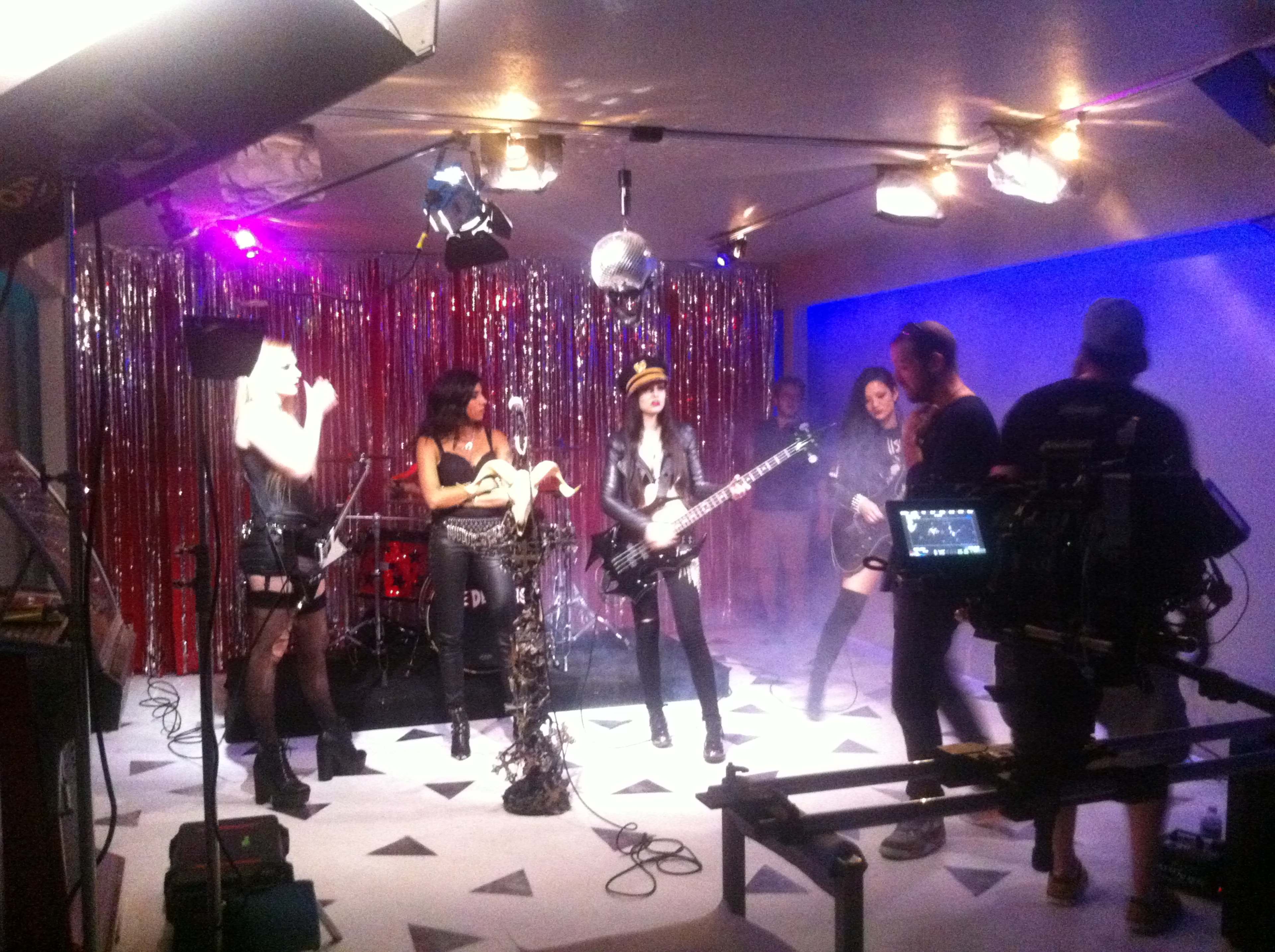 A band performs on a decorated stage with a disco ball and colorful lights, while a camera crew records the scene.