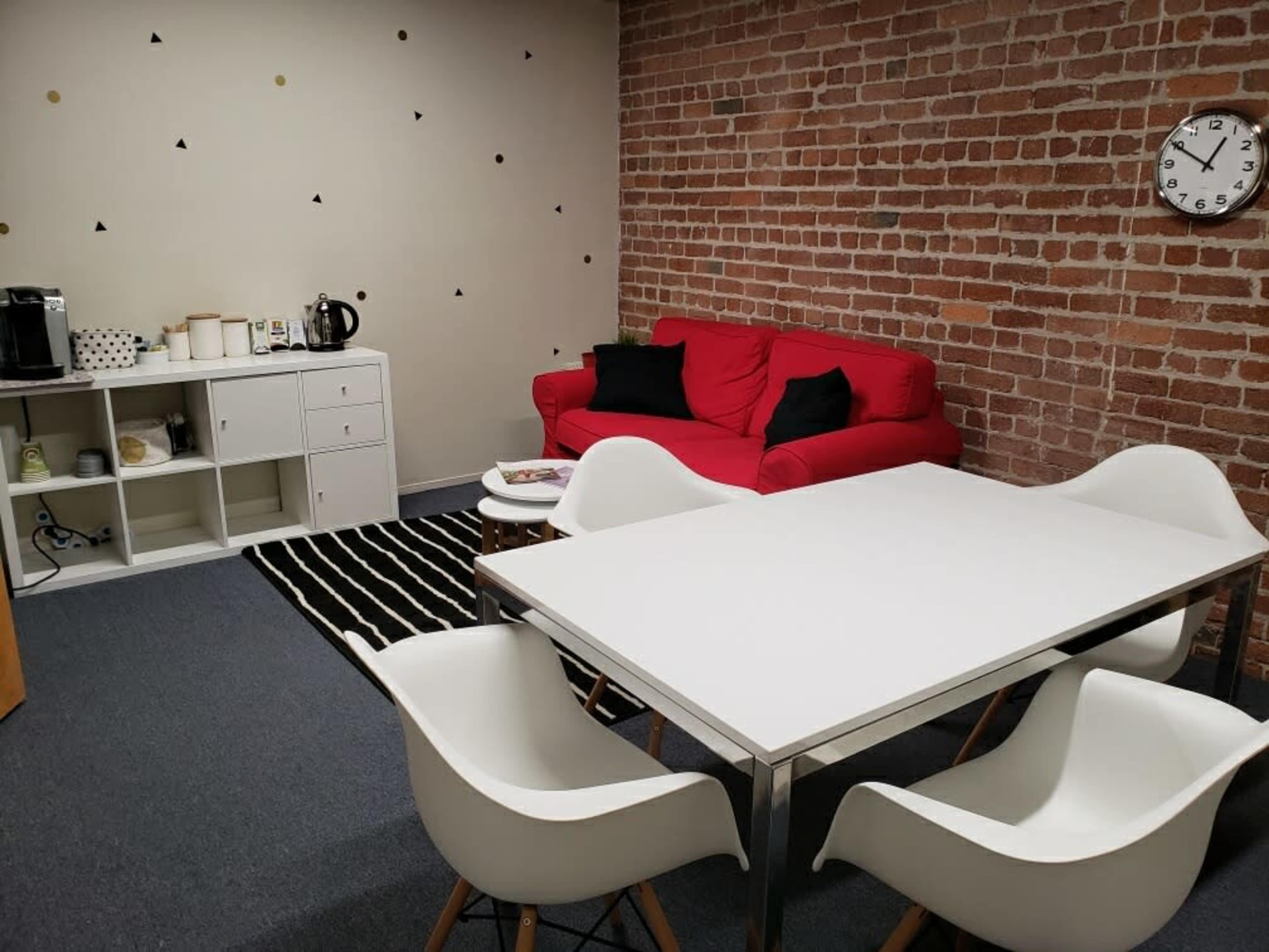A room with exposed brick walls, a red couch, a white dining table surrounded by four chairs, and a coffee station on a shelf.