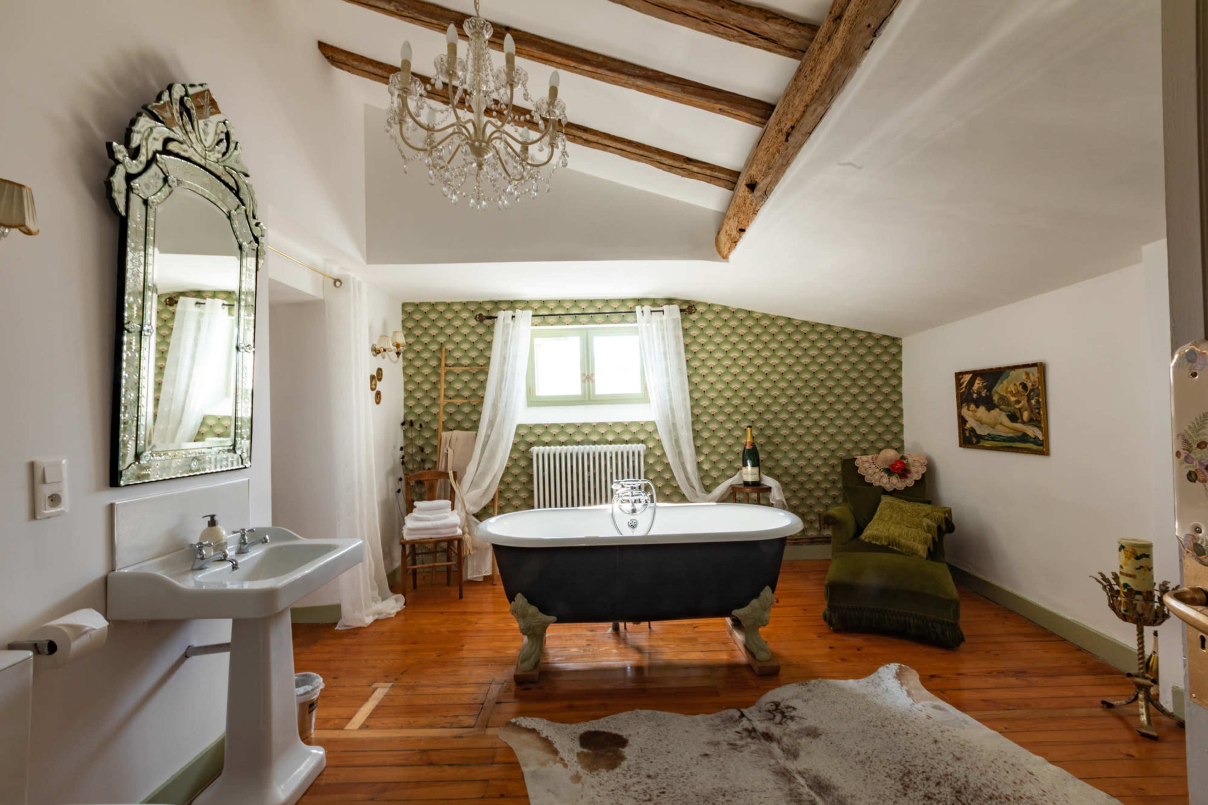 A spacious bathroom features a freestanding bathtub, a chandelier, and vintage decor with wooden beams and a patterned wall.