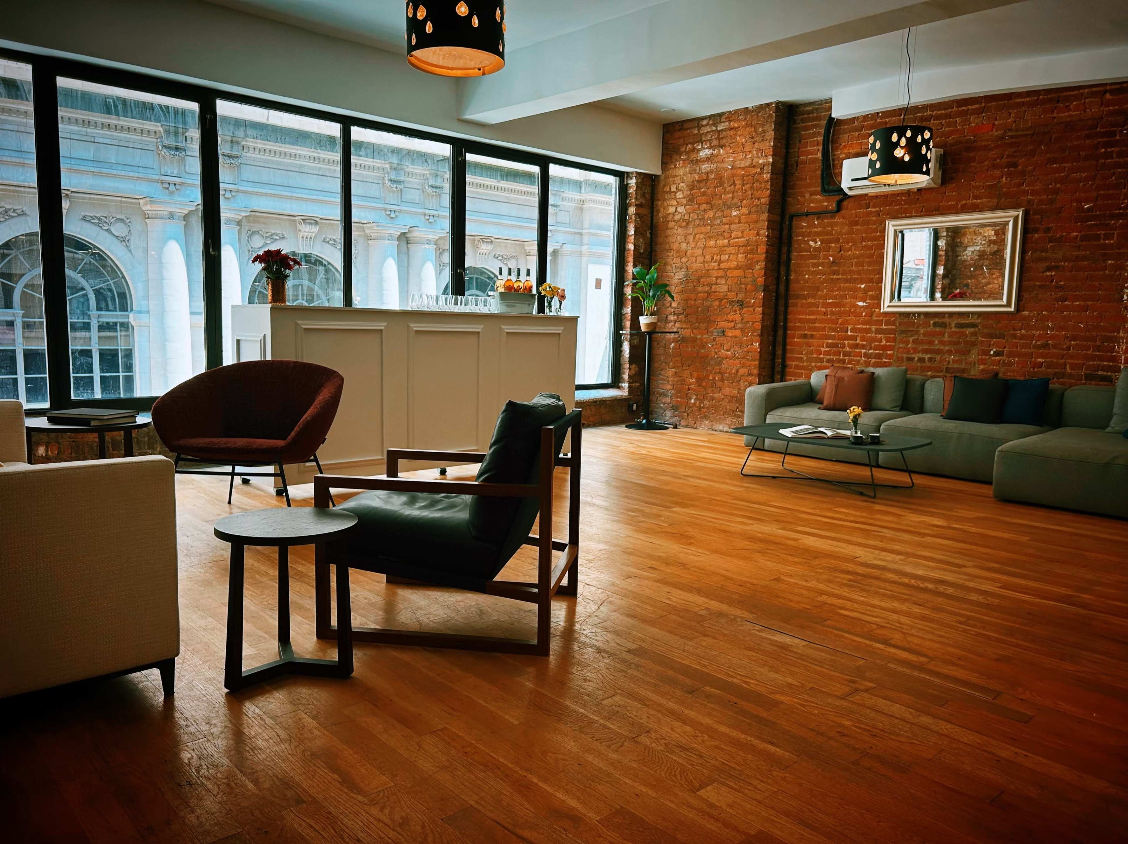 The scene features a modern, spacious room with large windows, exposed brick walls, and a mix of seating furniture arranged on wooden flooring.