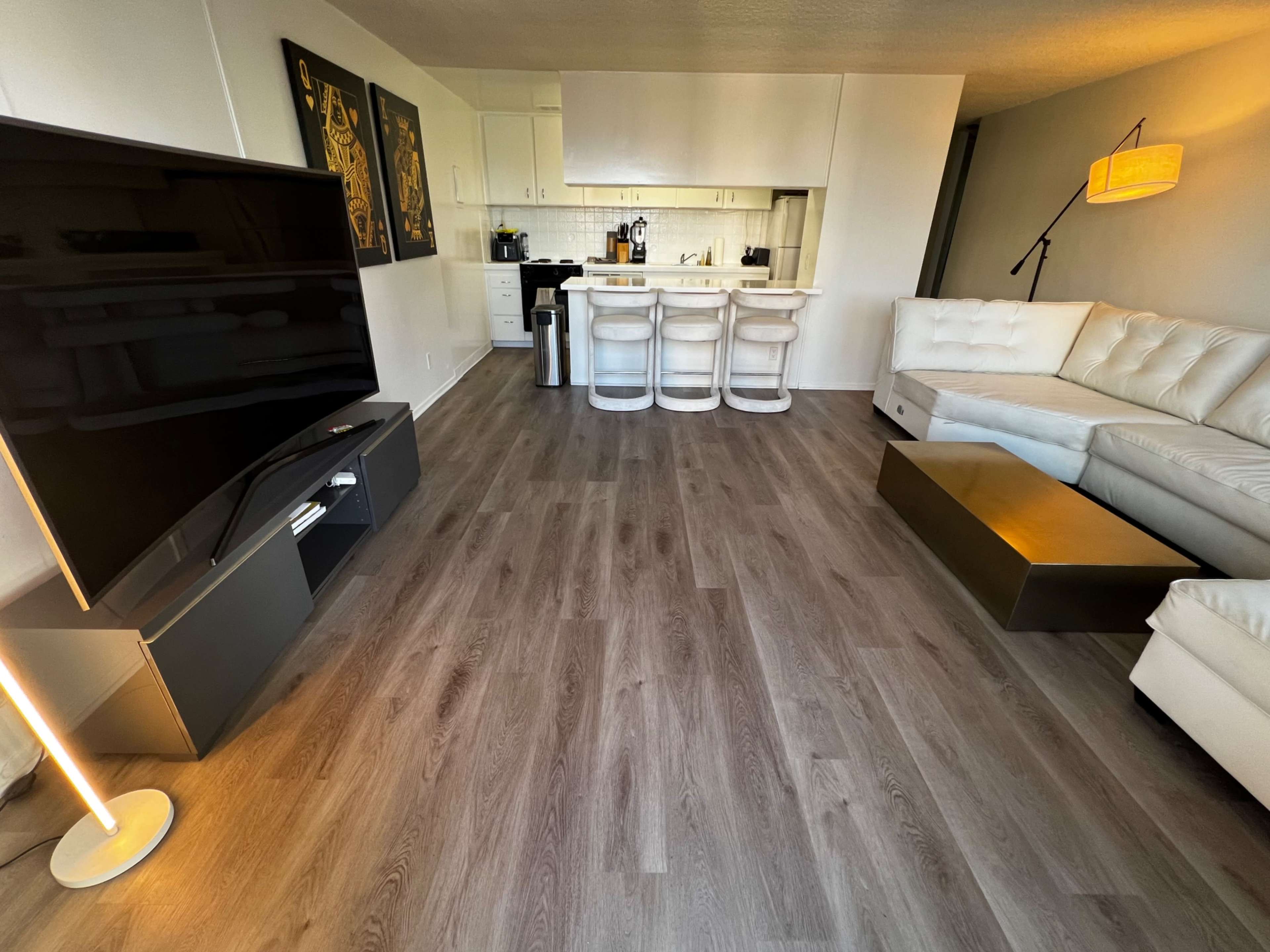 The image shows a modern living room with a large television, a white sectional sofa, a coffee table, and a kitchen area with bar stools in the background.