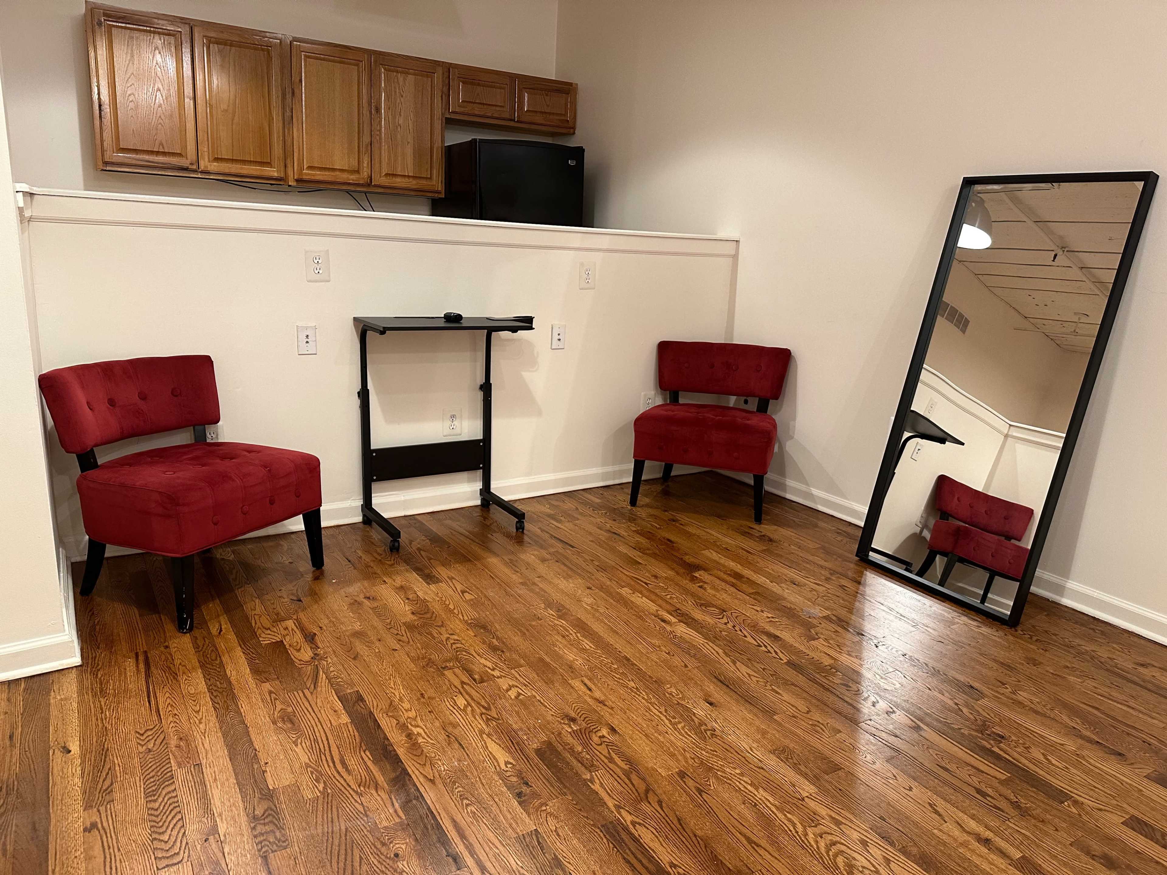 The image shows a room with two red upholstered chairs, a small black desk, and a large mirror against a light-colored wall, with wooden flooring and kitchen cabinetry in the background.