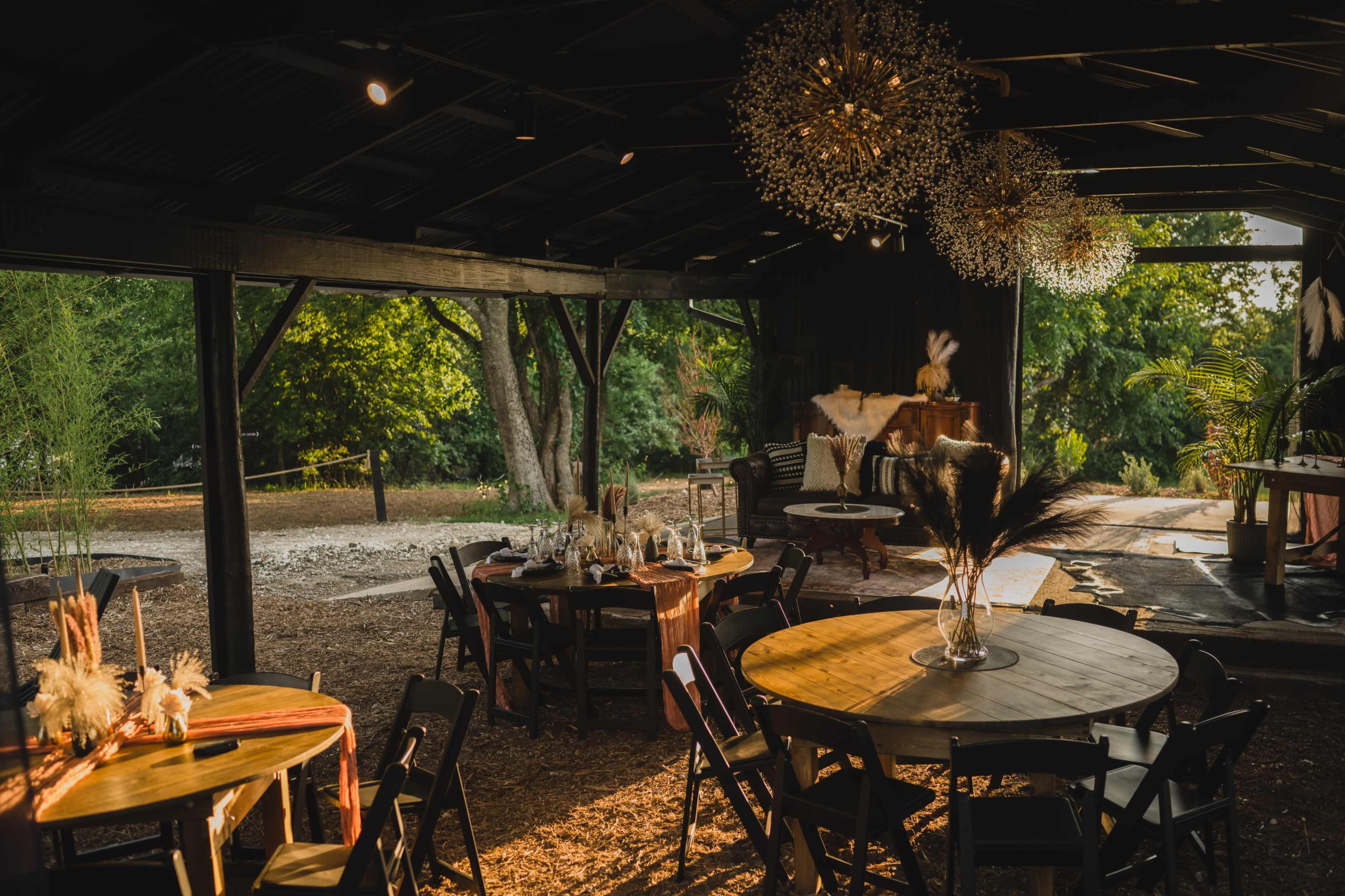The image shows a rustic indoor venue with round tables set for an event, illuminated by decorative chandeliers and surrounded by greenery.