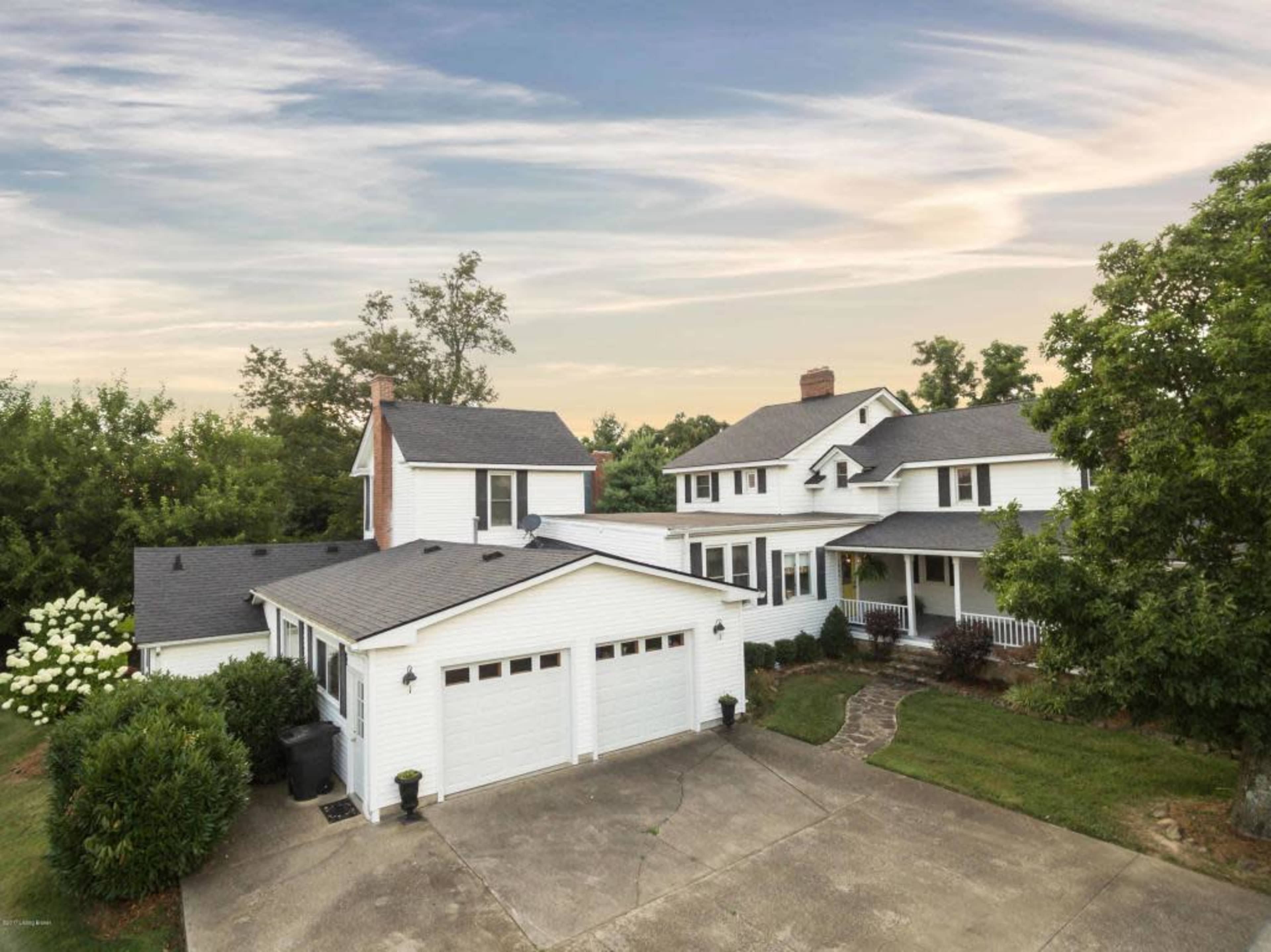 A white two-story house with multiple sections and a garage is surrounded by greenery and features a paved driveway.