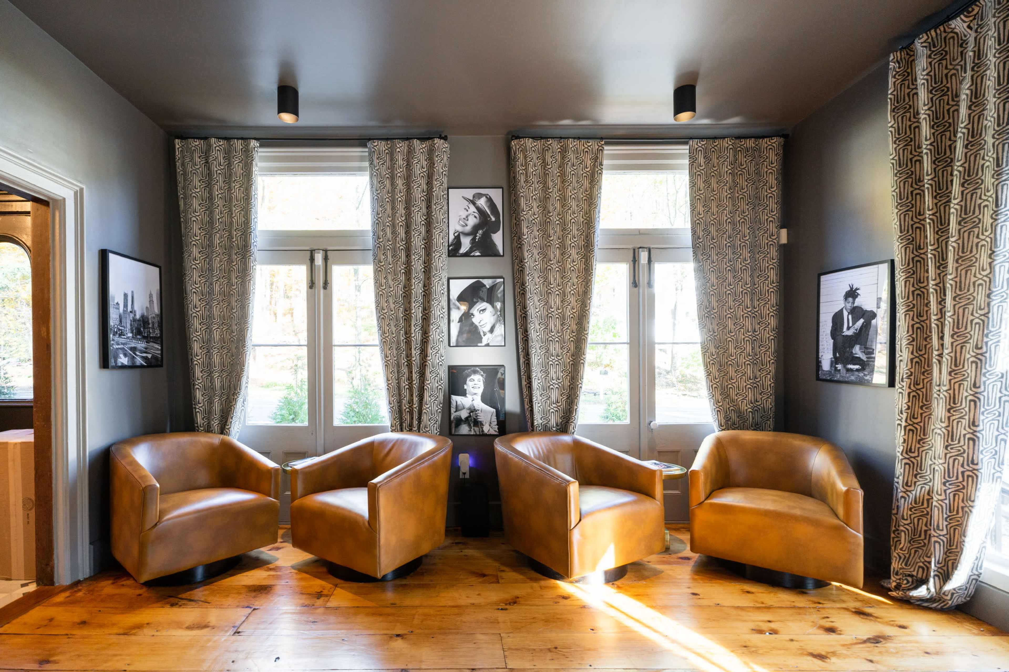 The image shows a cozy seating area with three brown chairs arranged facing each other, surrounded by large windows and framed black-and-white photographs on the walls.