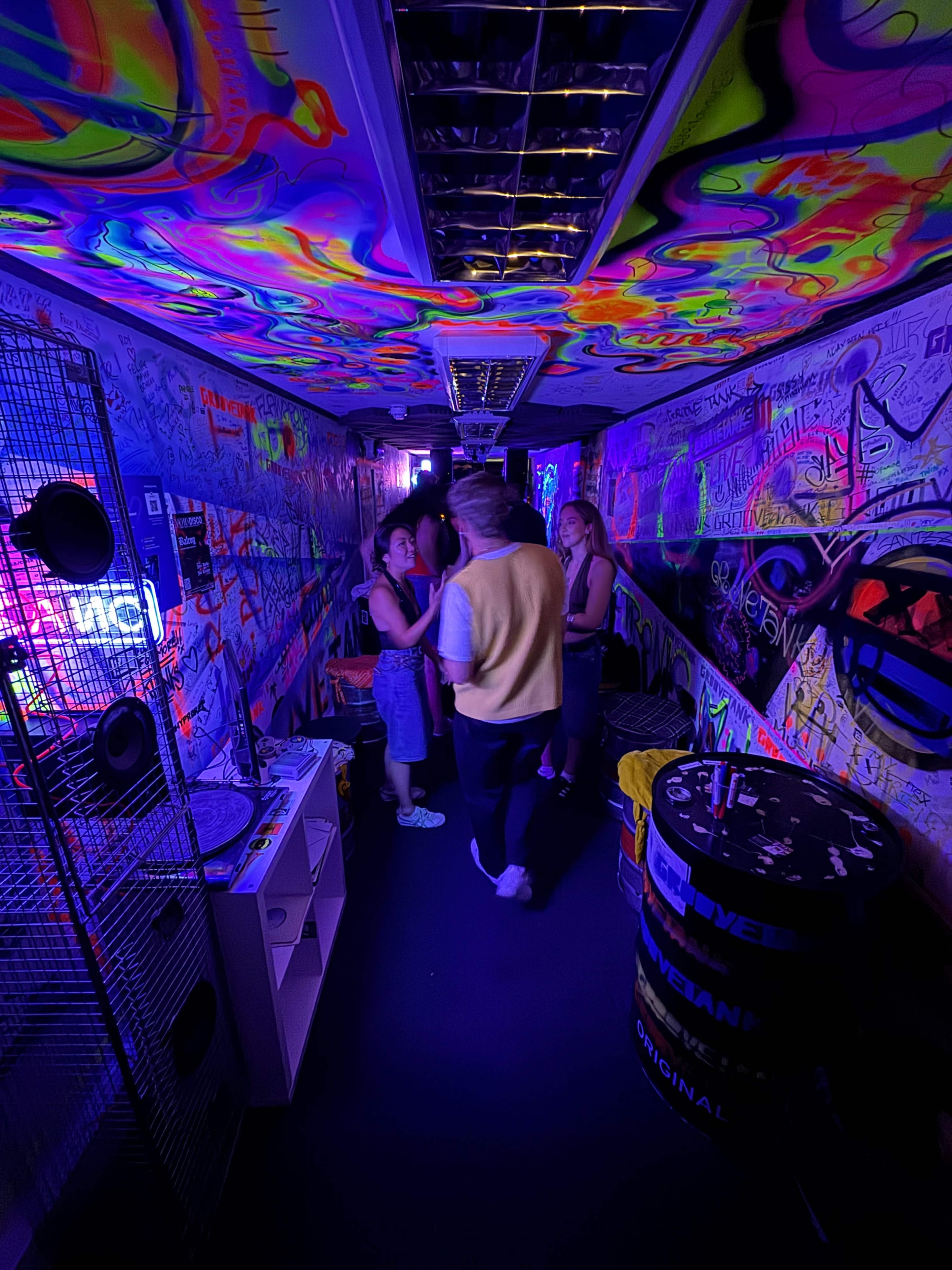 A group of people stands in a brightly colored hallway adorned with graffiti and illuminated by neon lights.