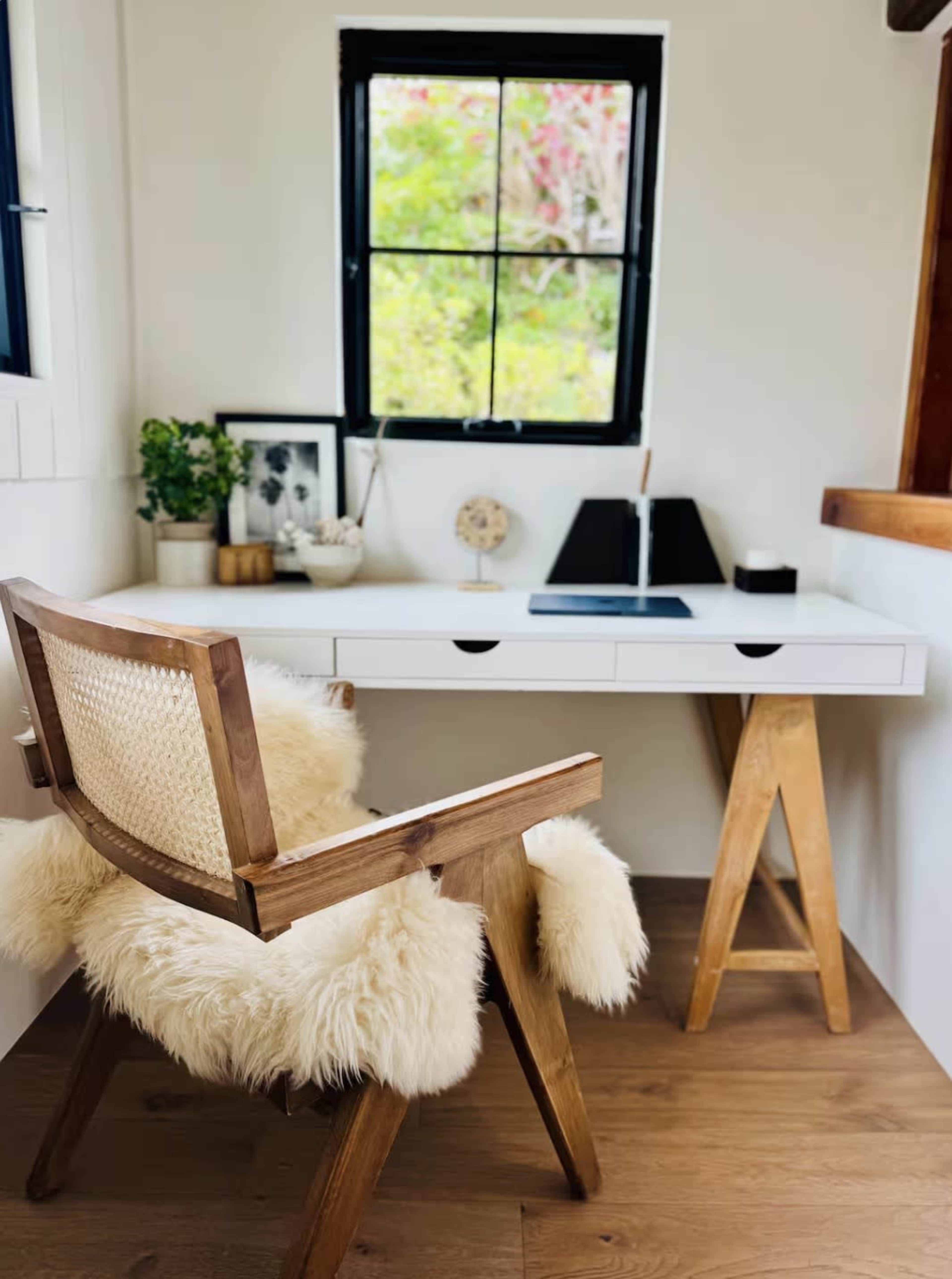 The image shows a minimalist desk area with a white desk, a wooden chair, and a window overlooking greenery.
