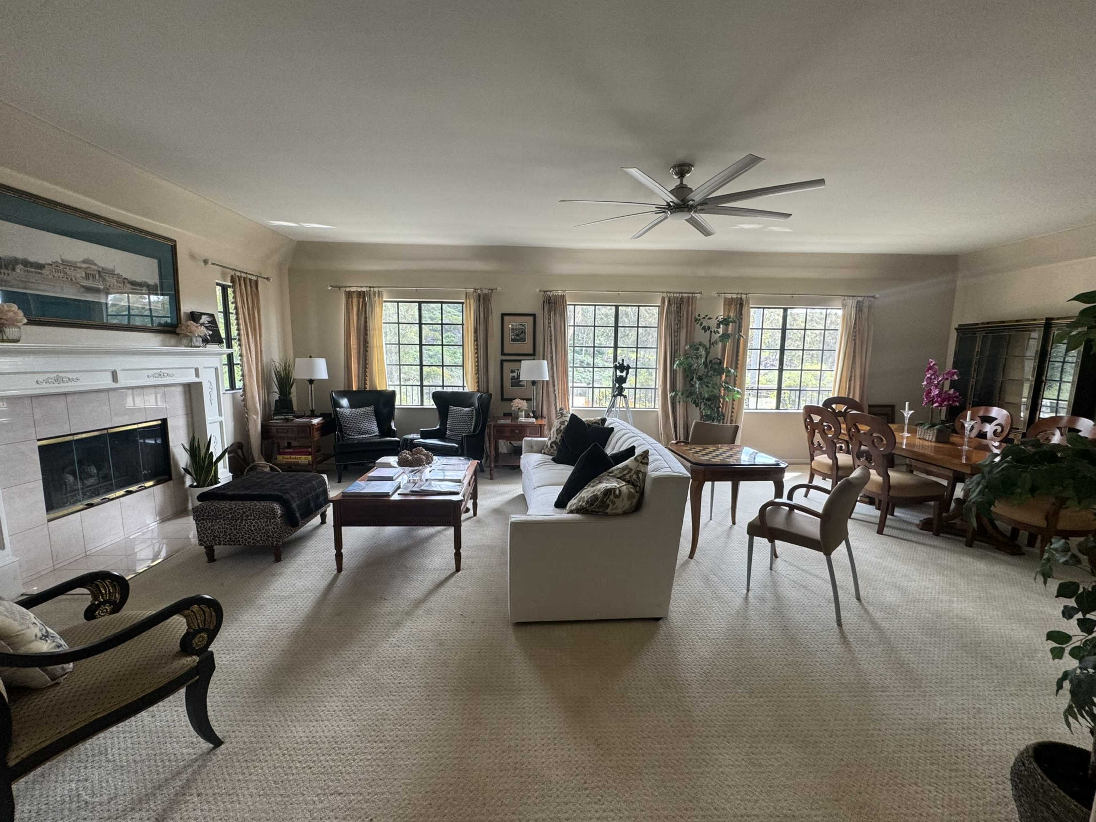 A spacious living room with a mix of seating arrangements, including a white sofa, chairs, and a dining table, all surrounded by large windows and decorative plants.