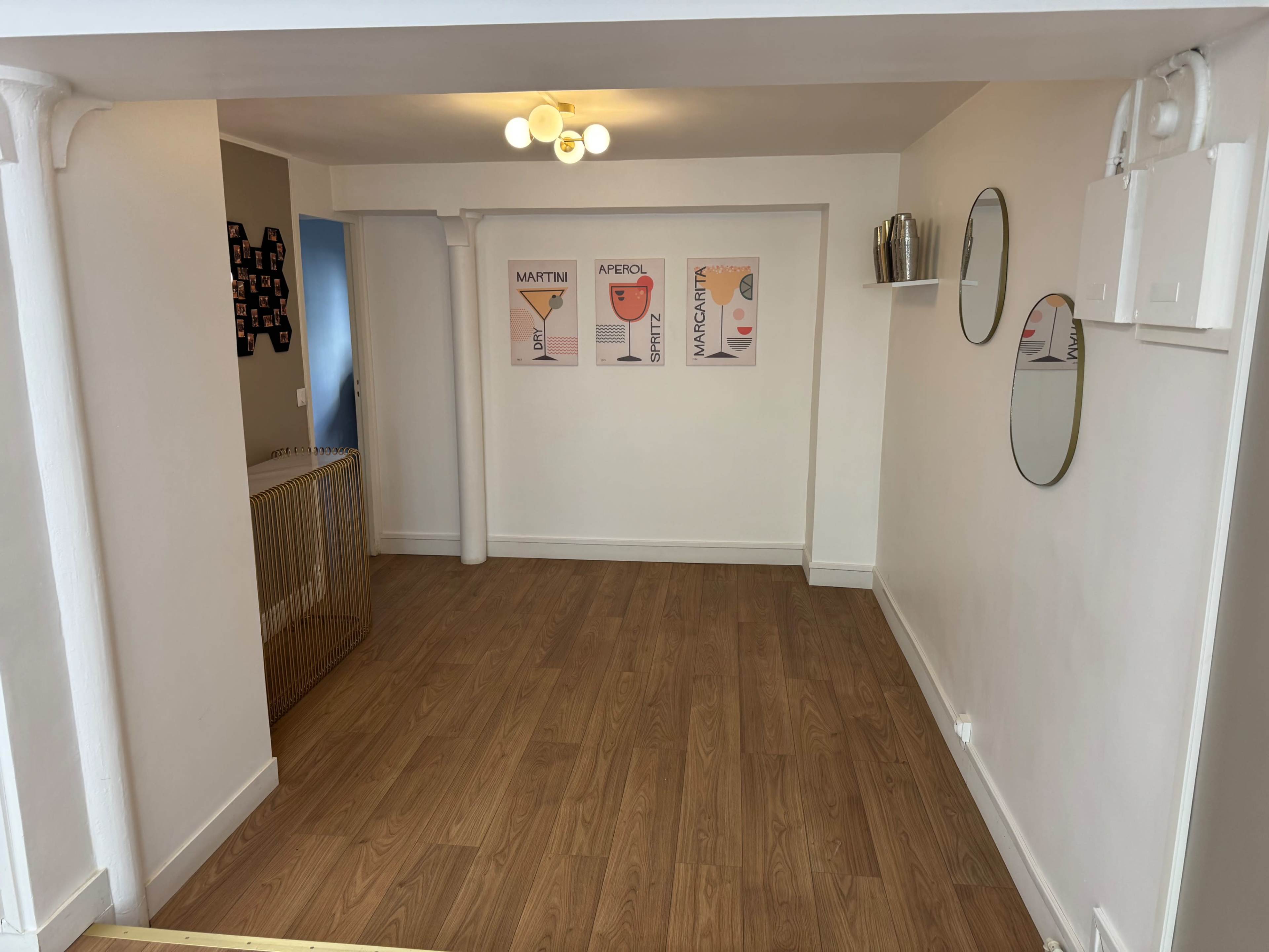 The image shows a minimalist interior space with wooden flooring, featuring light-colored walls, framed art prints, and decorative mirrors.