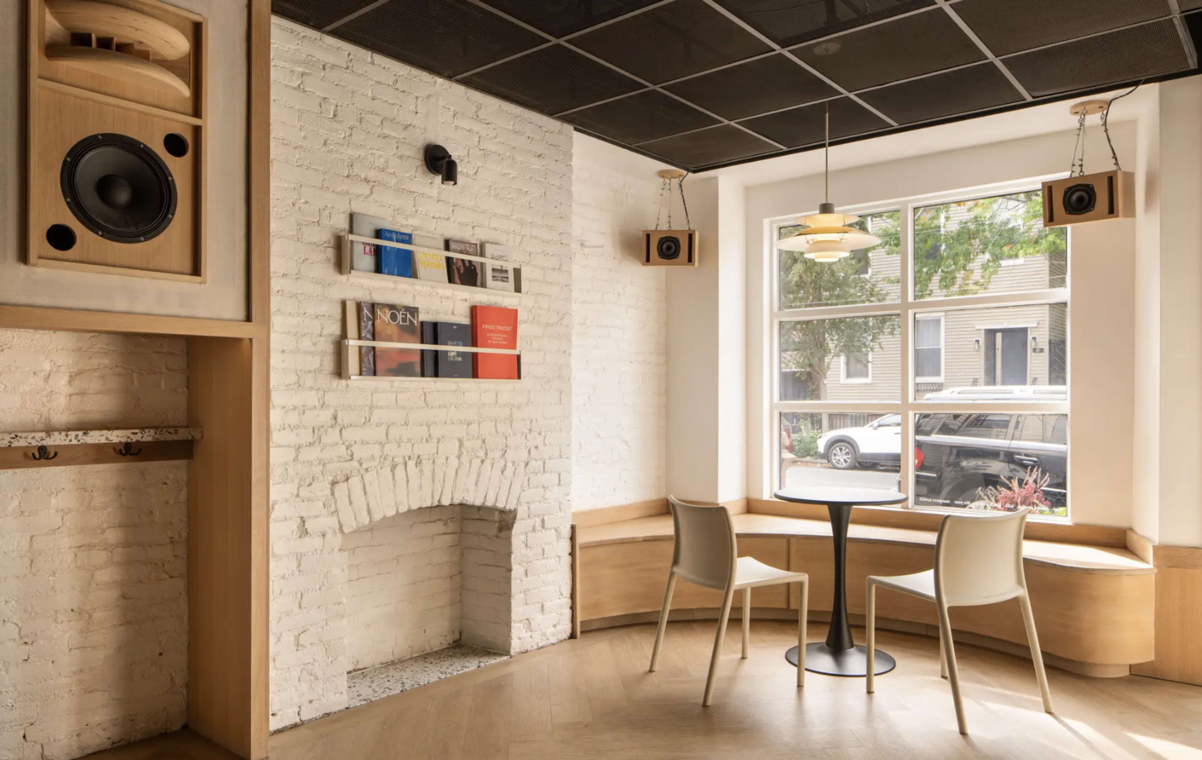 The image shows a minimalist café interior featuring a circular table with two chairs, a brick wall with books displayed, and large windows providing natural light.