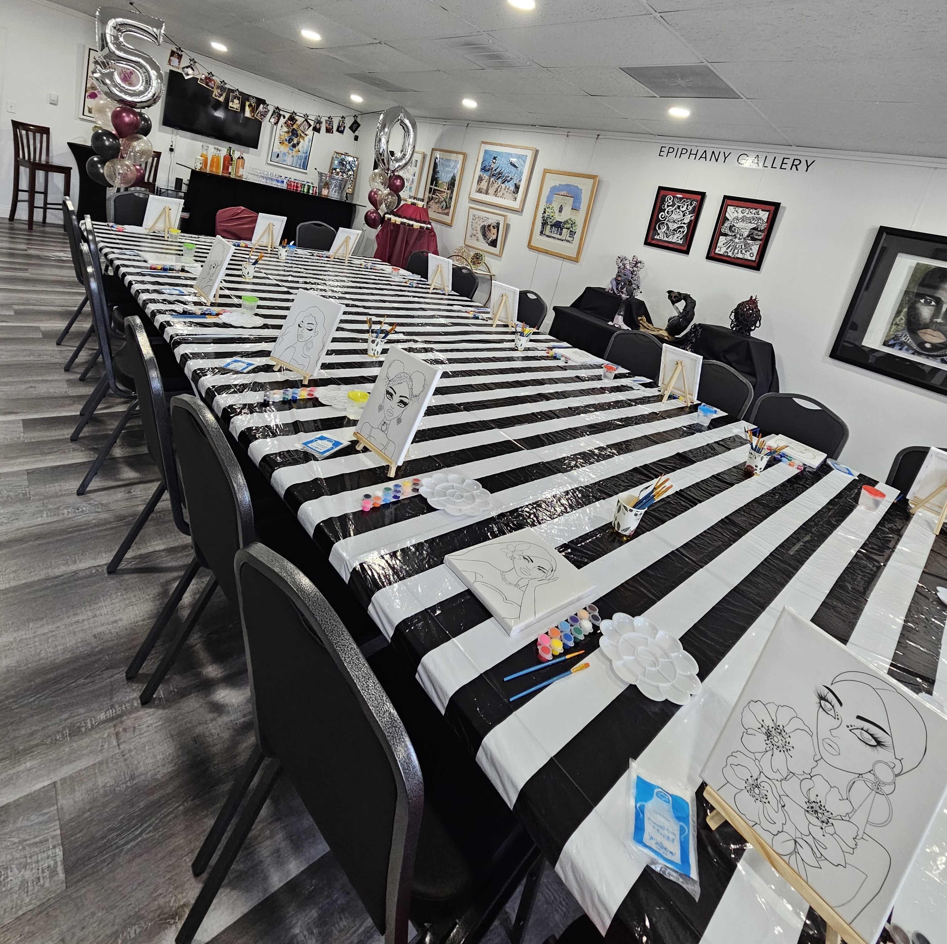 A long table covered with a black and white striped tablecloth is set up in a gallery with art supplies and canvases ready for a painting event.
