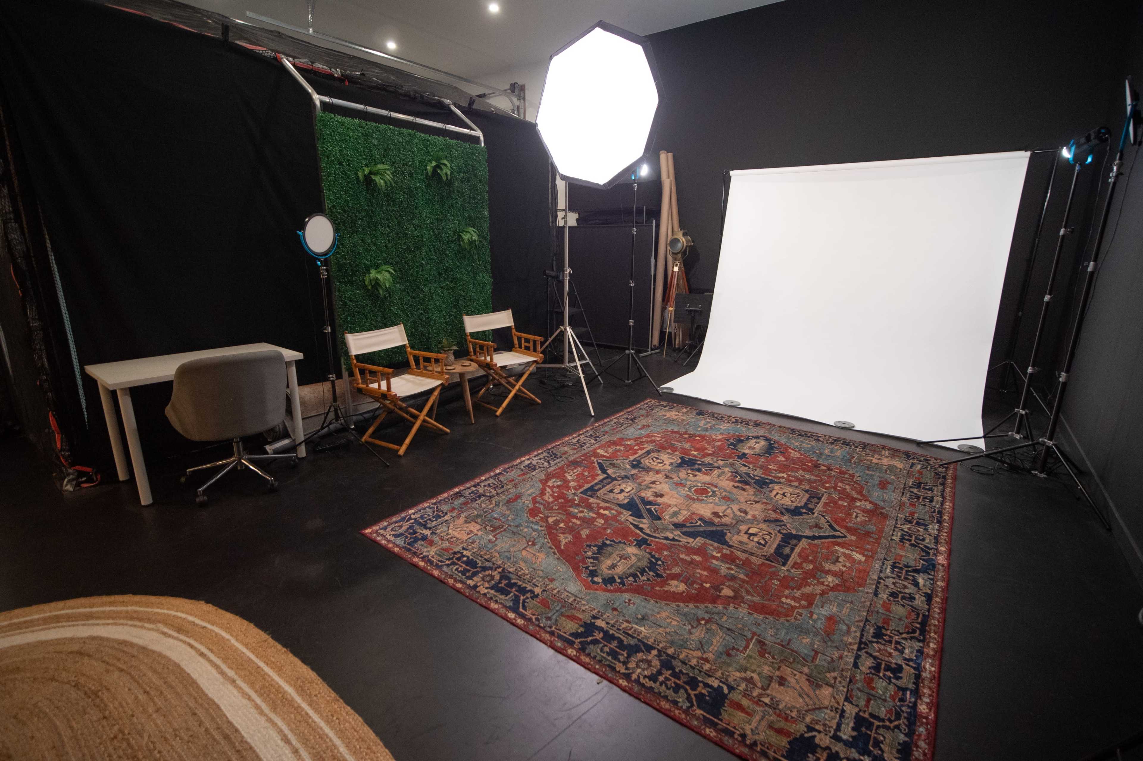 The image shows a photography studio with a white backdrop, a decorative rug on the floor, chairs, and lighting equipment in the setup.