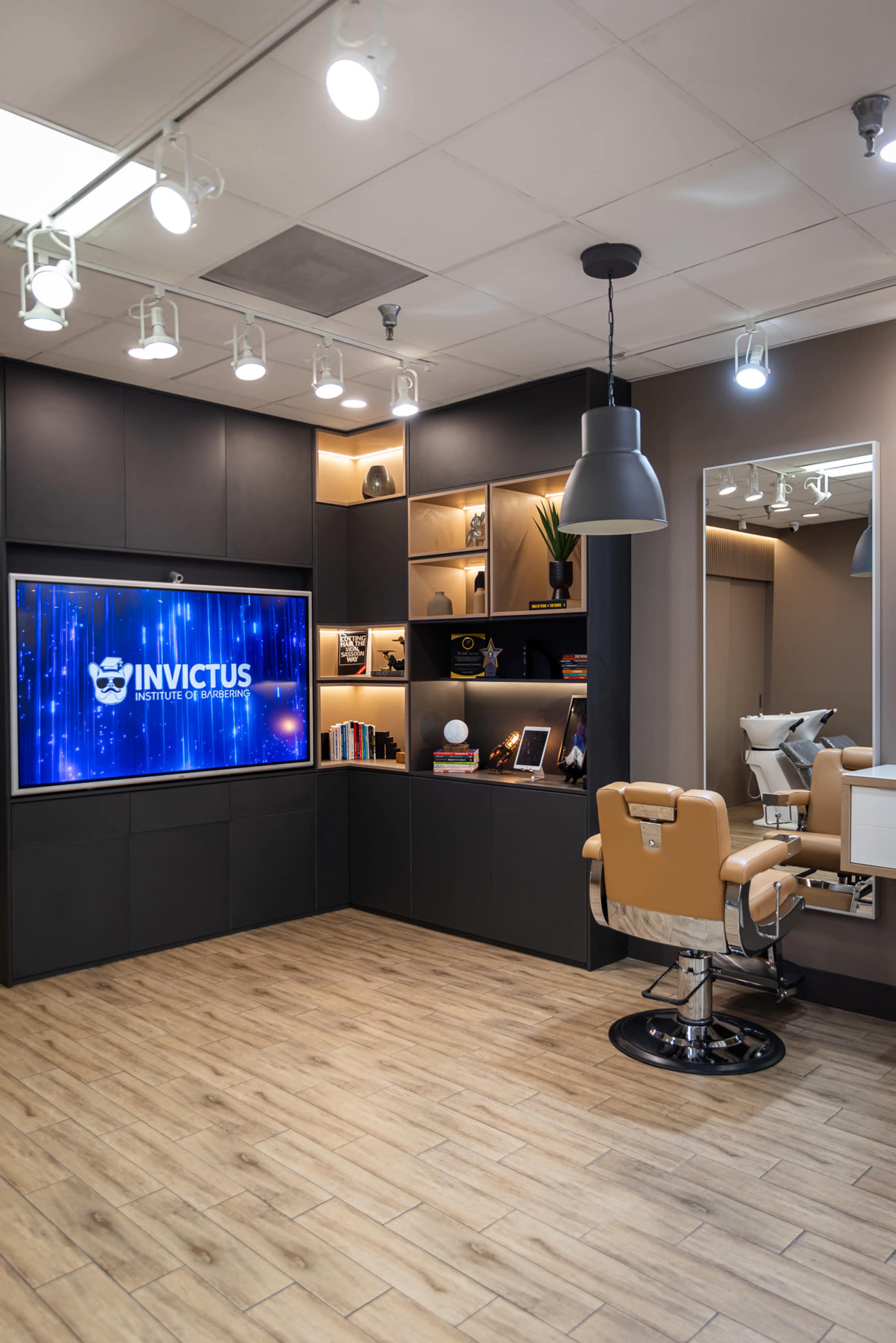 The image shows a modern barbershop interior featuring a large screen displaying the name "INVICTUS," a stylish chair, and shelves with decor and books.