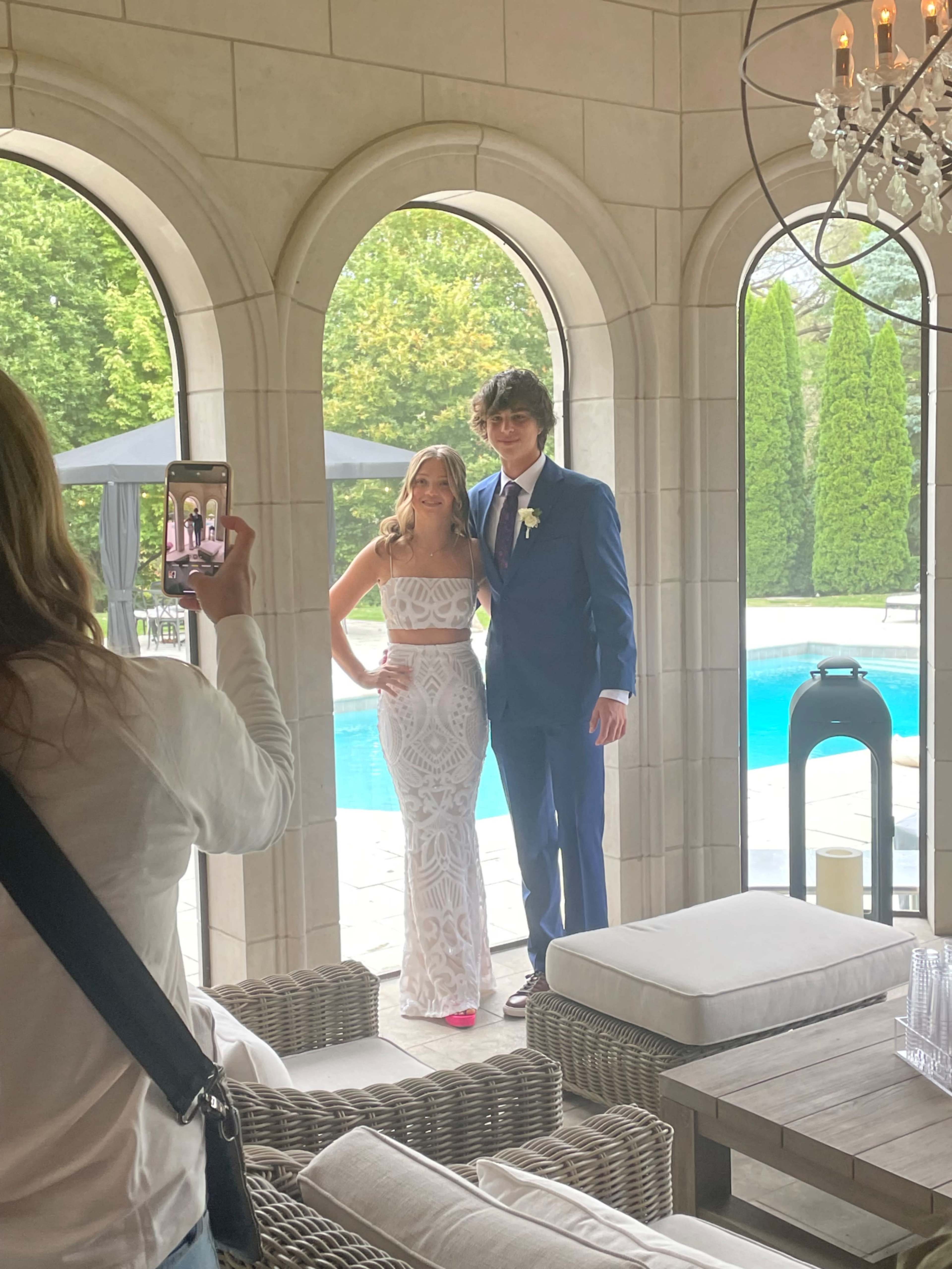 A young woman in a white dress poses with a man in a blue suit near a poolside, while another person takes their picture.