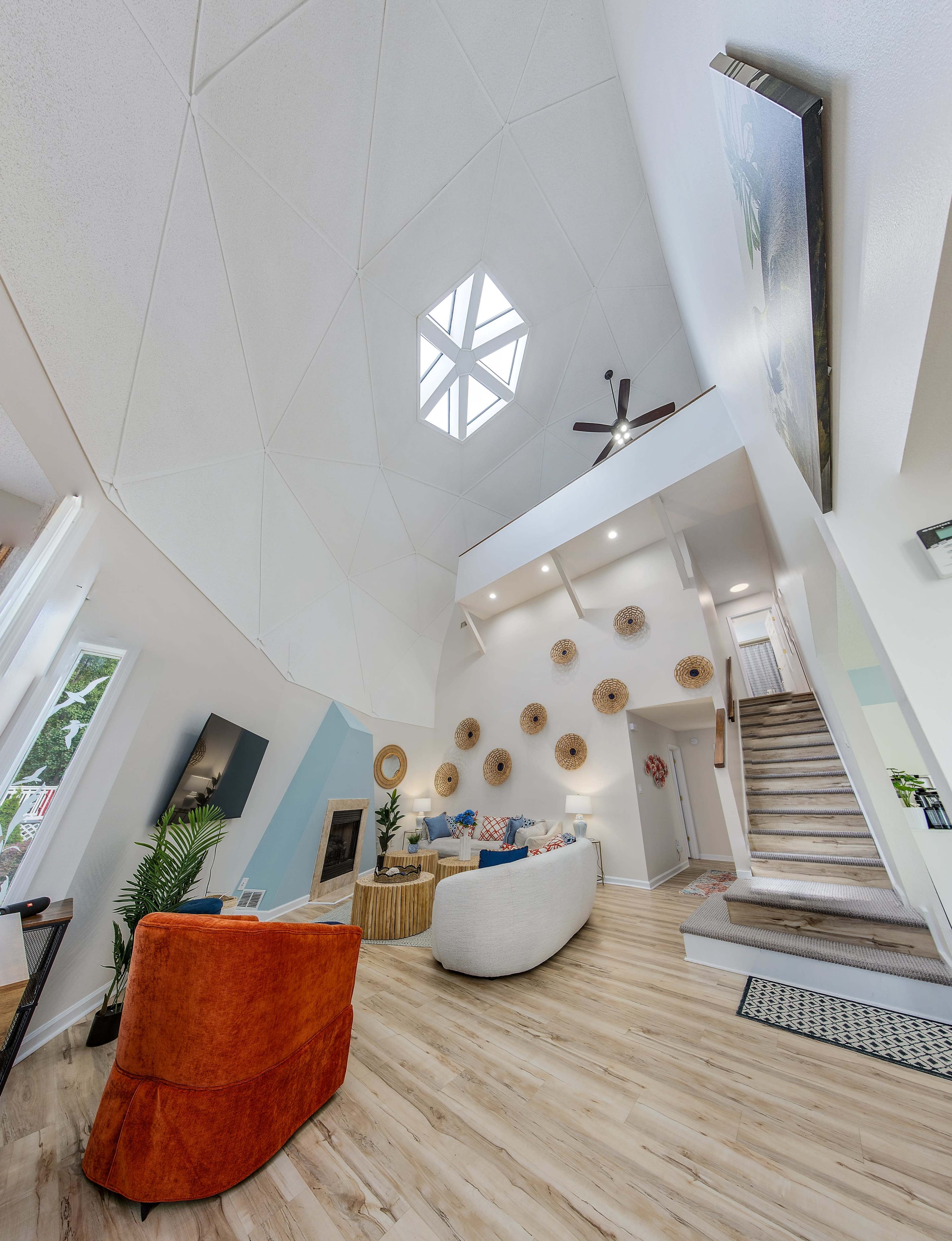 The image shows a spacious, modern living room with a high, geometric ceiling and a staircase leading to an upper level.