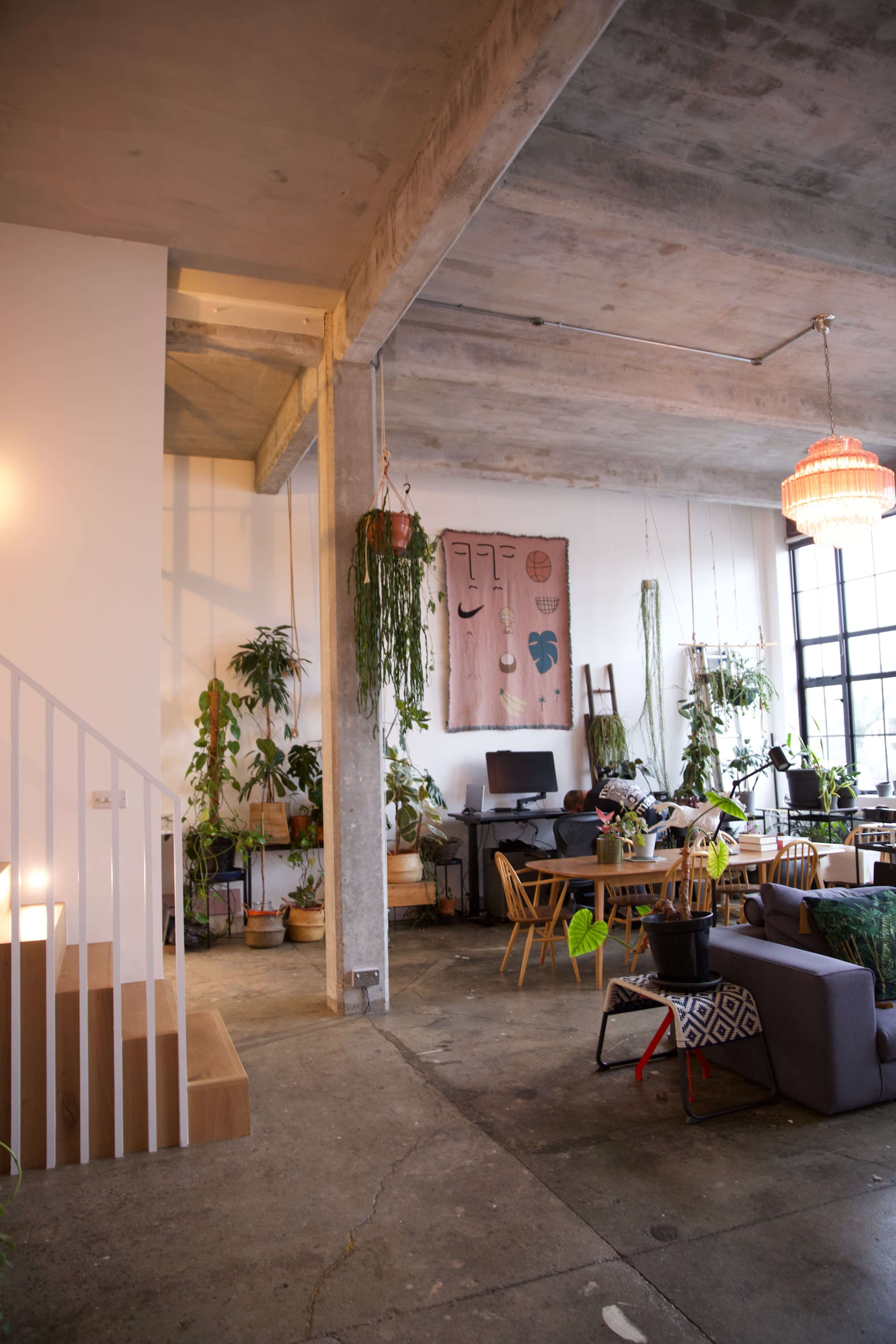 The image depicts a modern, open-concept living space with concrete ceilings, plants decoratively arranged around the room, and a mix of furniture including a dining table and seating area.