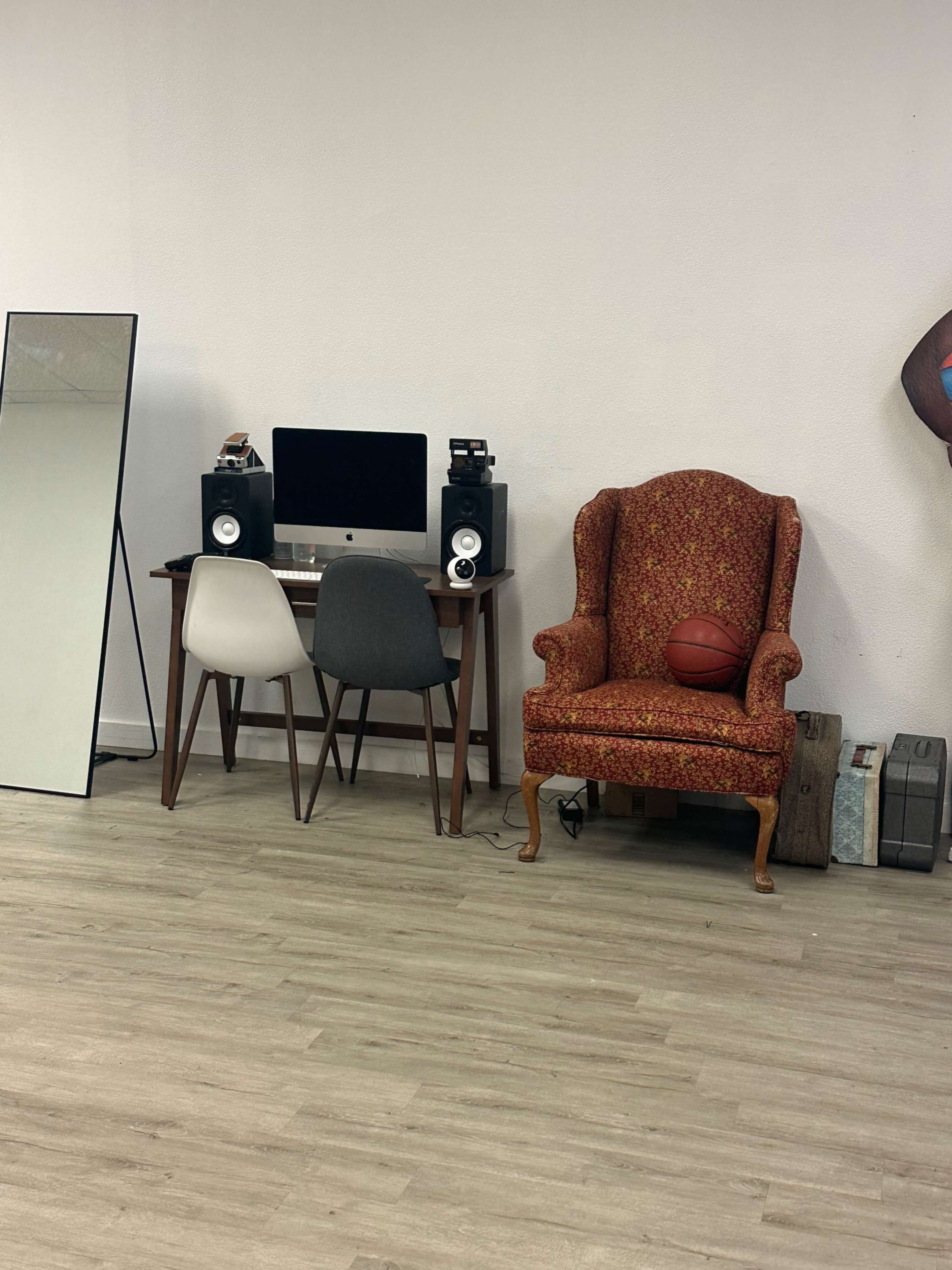 A workspace featuring a desk with a computer and speakers, flanked by two chairs and a basketball resting on an ornate armchair, alongside a mirror and a few storage boxes.