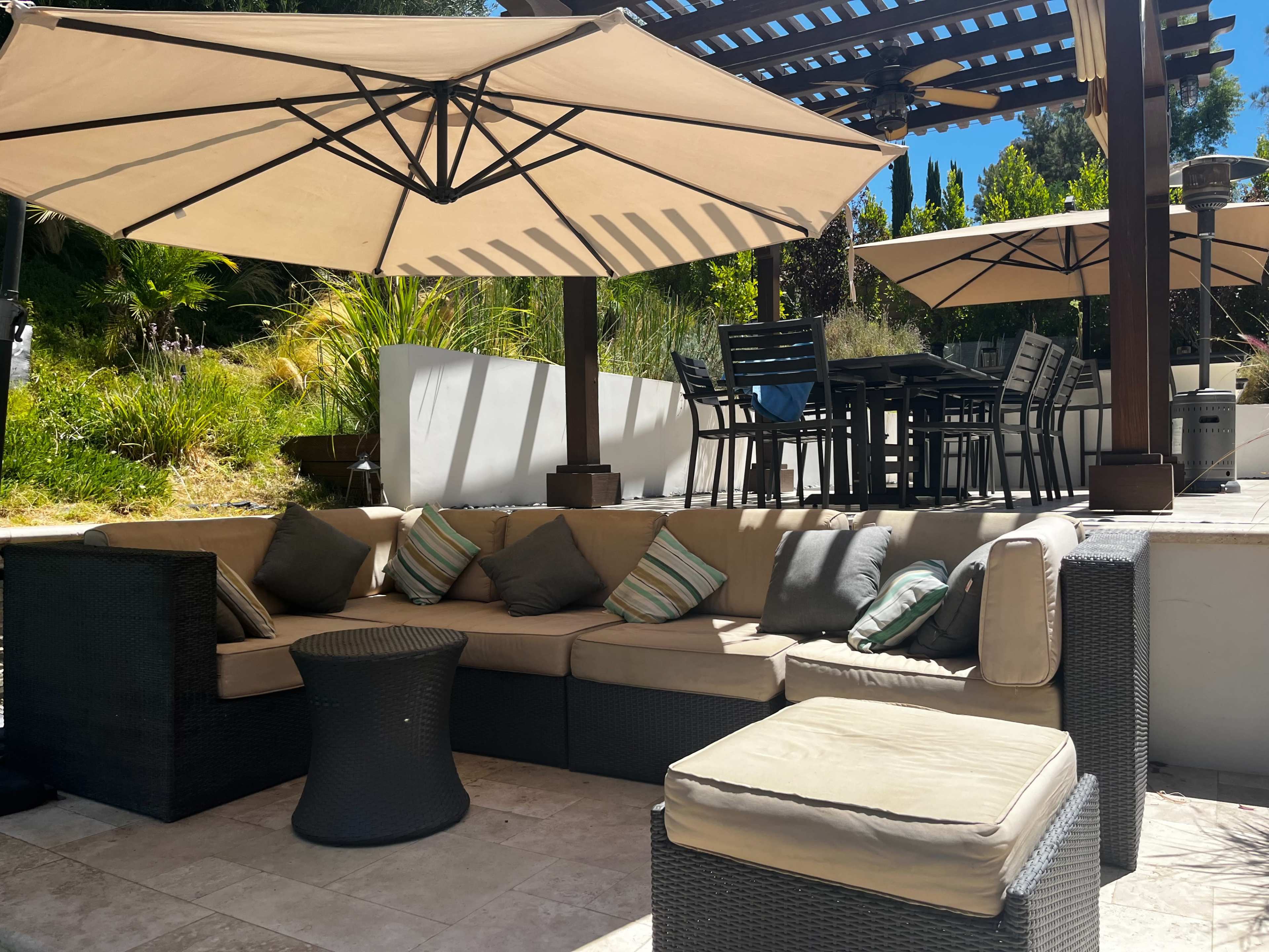 A shaded outdoor seating area features a sectional sofa with decorative cushions, a small round table, and a dining set under large umbrellas.