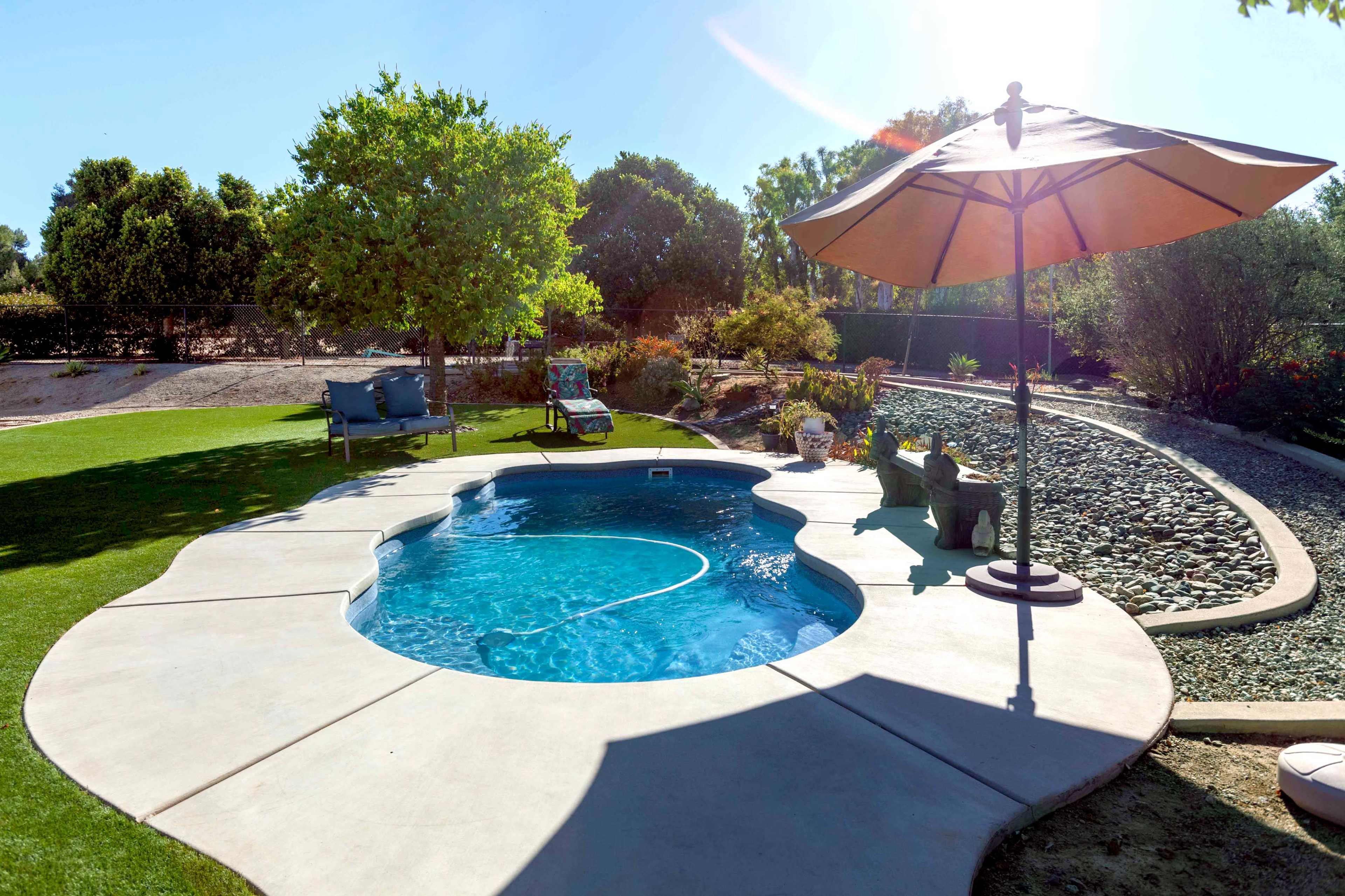 A small, circular pool is surrounded by a concrete patio, lawn, and a lounge chair under an umbrella, with a landscaped garden in the background.