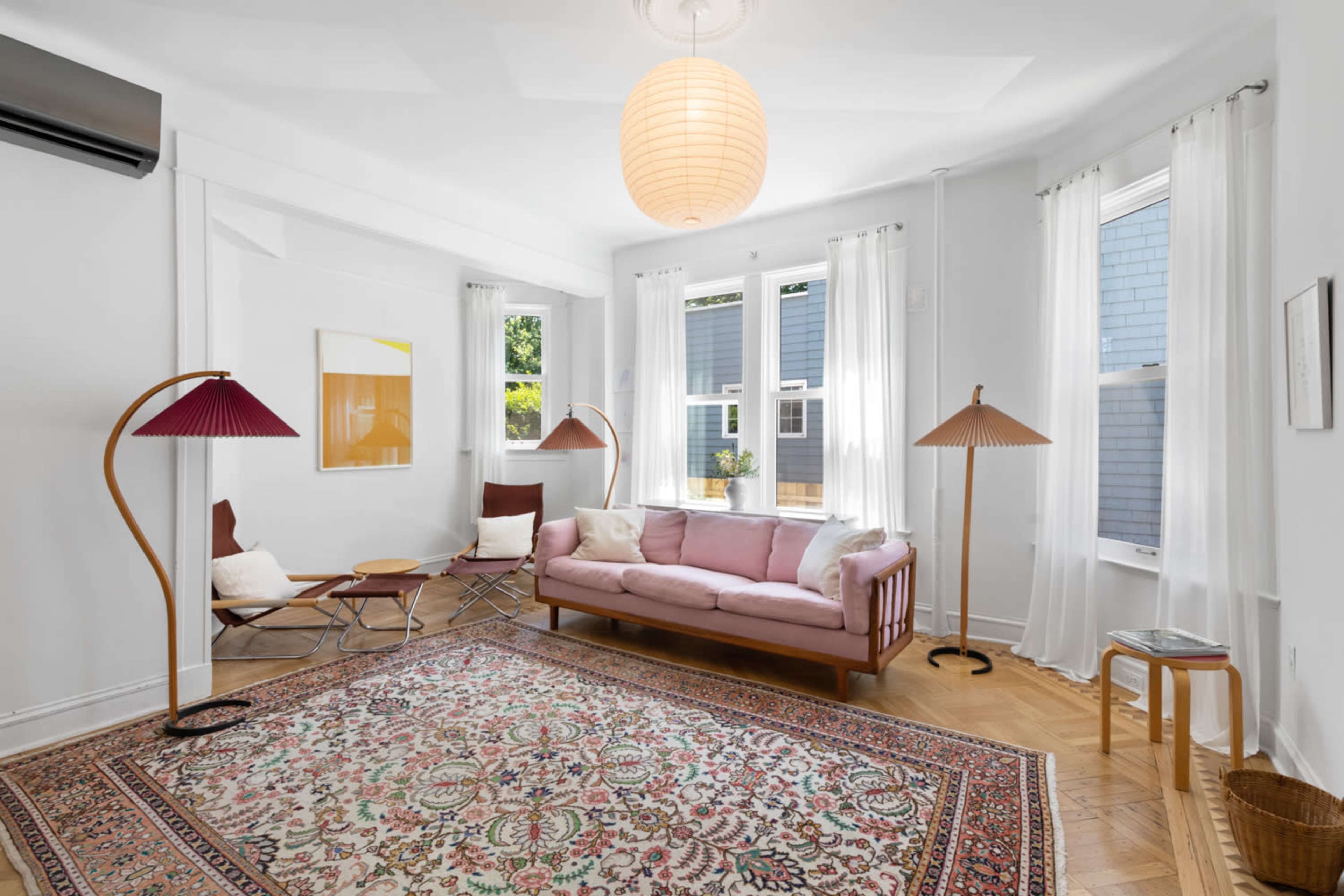 The image shows a bright living room with a pink sofa, two unique floor lamps, a large area rug, and several windows allowing natural light.
