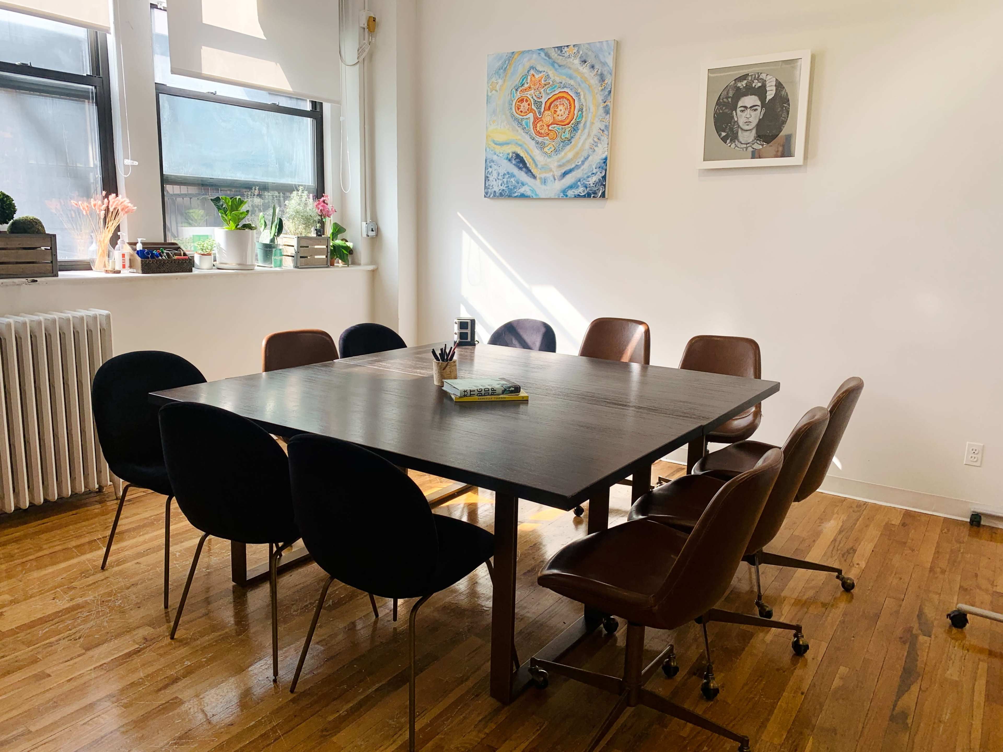 A wooden table surrounded by eight office chairs sits in a bright room with large windows and a decorated wall featuring artwork.