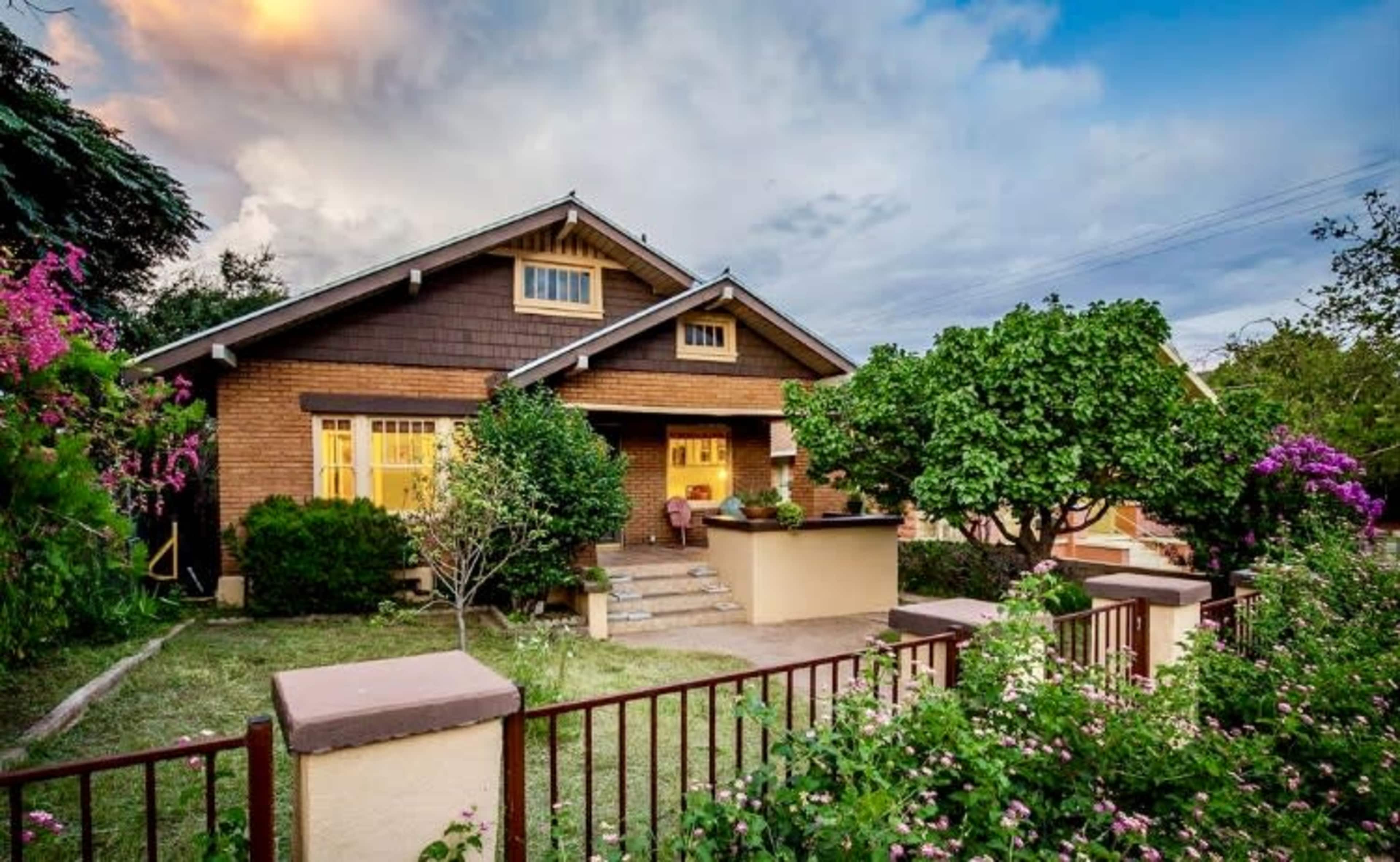 A brick bungalow with a gabled roof is surrounded by green shrubs and colorful flowers in a residential setting.
