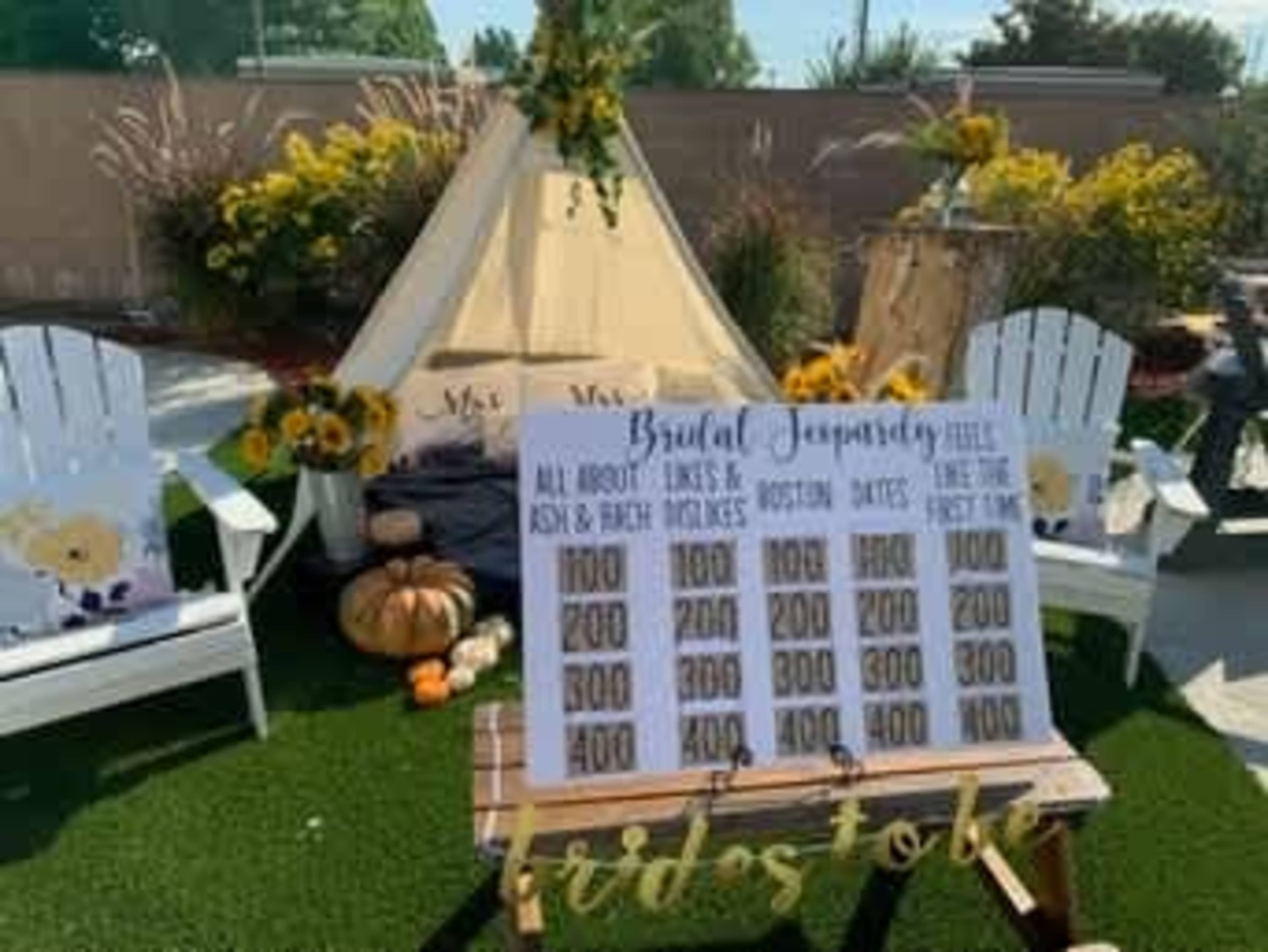 The image features a backyard scene with a decorative tent, two white adirondack chairs, sunflowers, a pumpkin, and a sign displaying "Brutal Jeopardy" with various point values for a game.