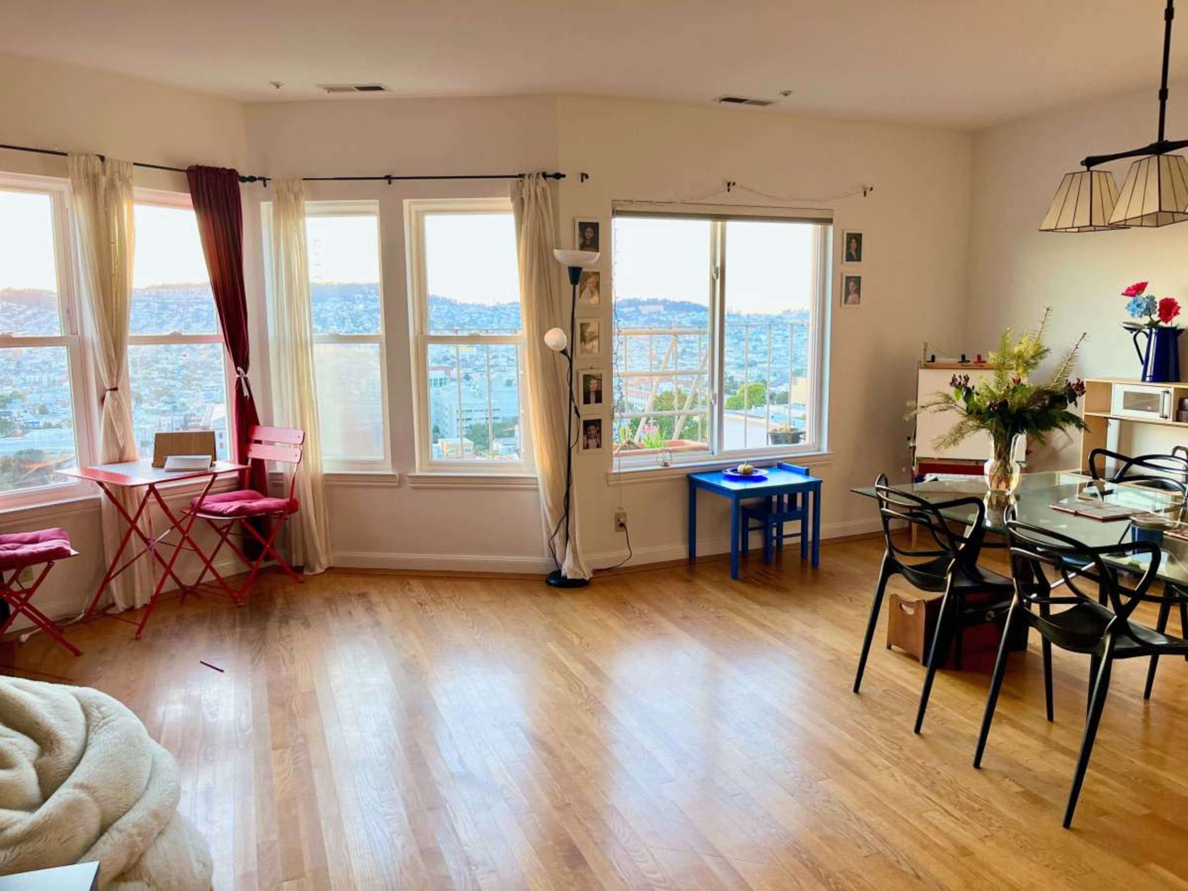 The image shows a bright room with large windows overlooking a cityscape, featuring a dining table, chairs, and a small seating area.
