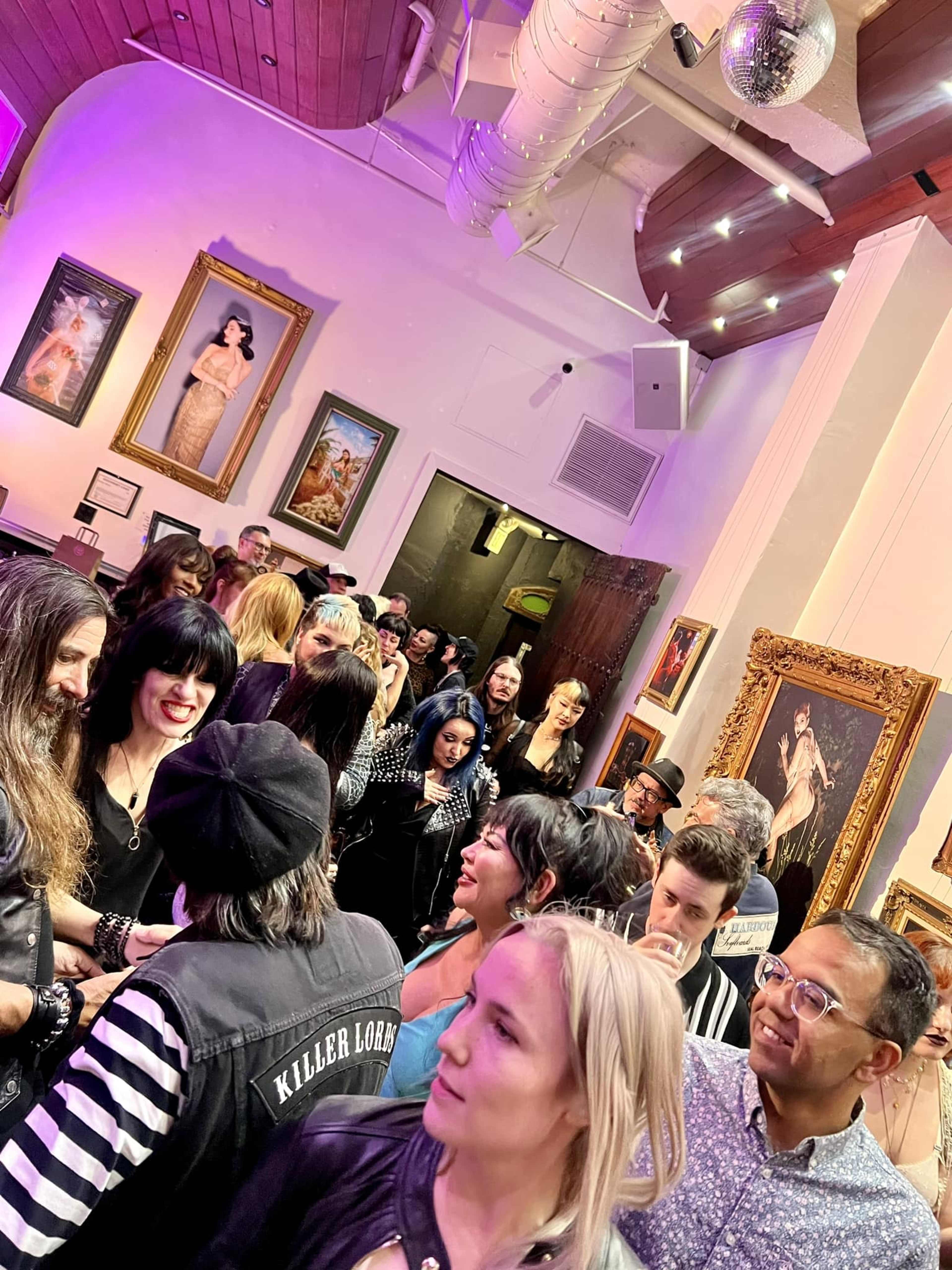 A crowded art gallery features people mingling among framed paintings, with a large disco ball hanging from the ceiling.