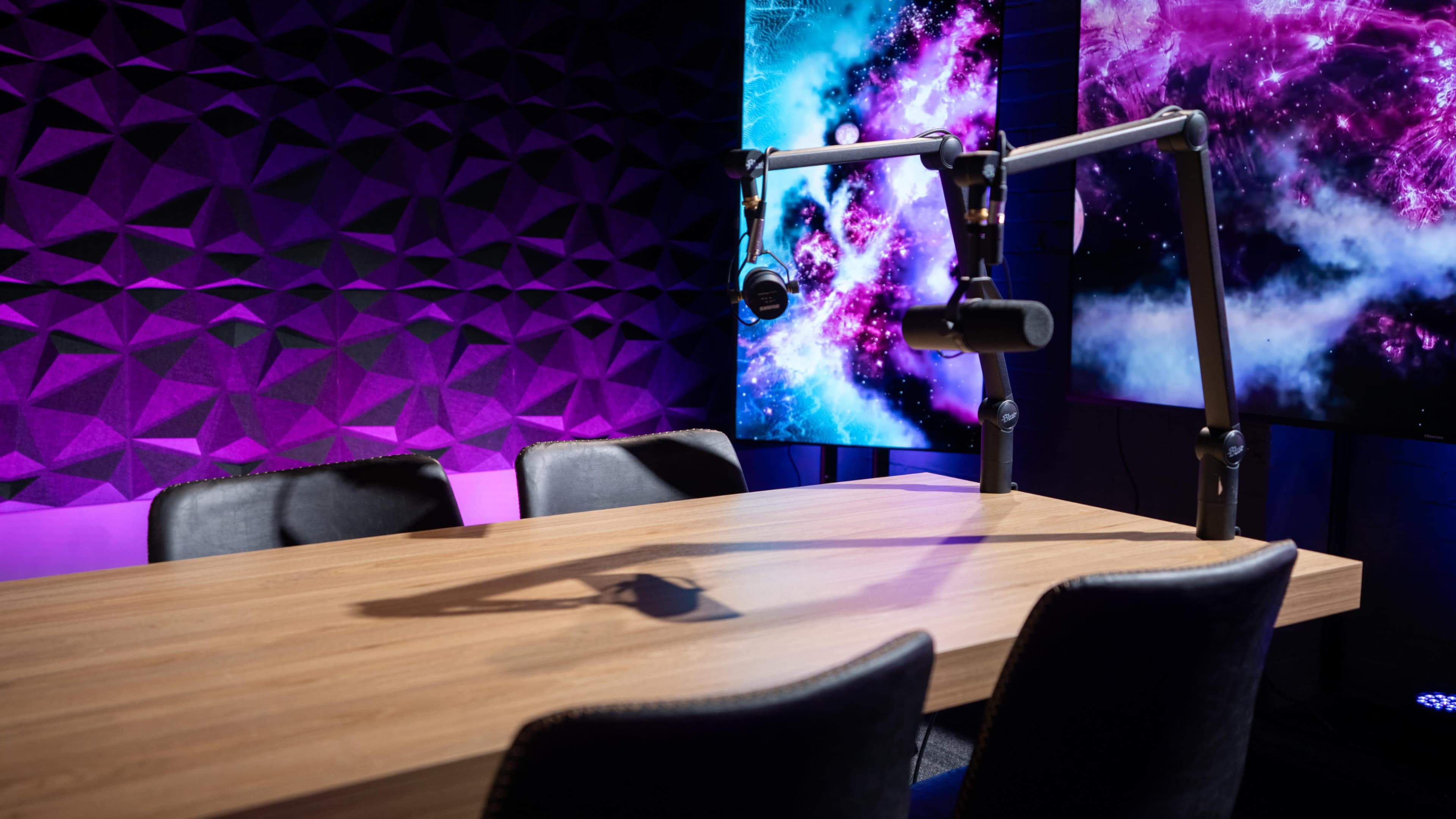 A podcast studio features a wooden table with black chairs and microphone stands, set against a colorful backdrop of purple and blue graphics.