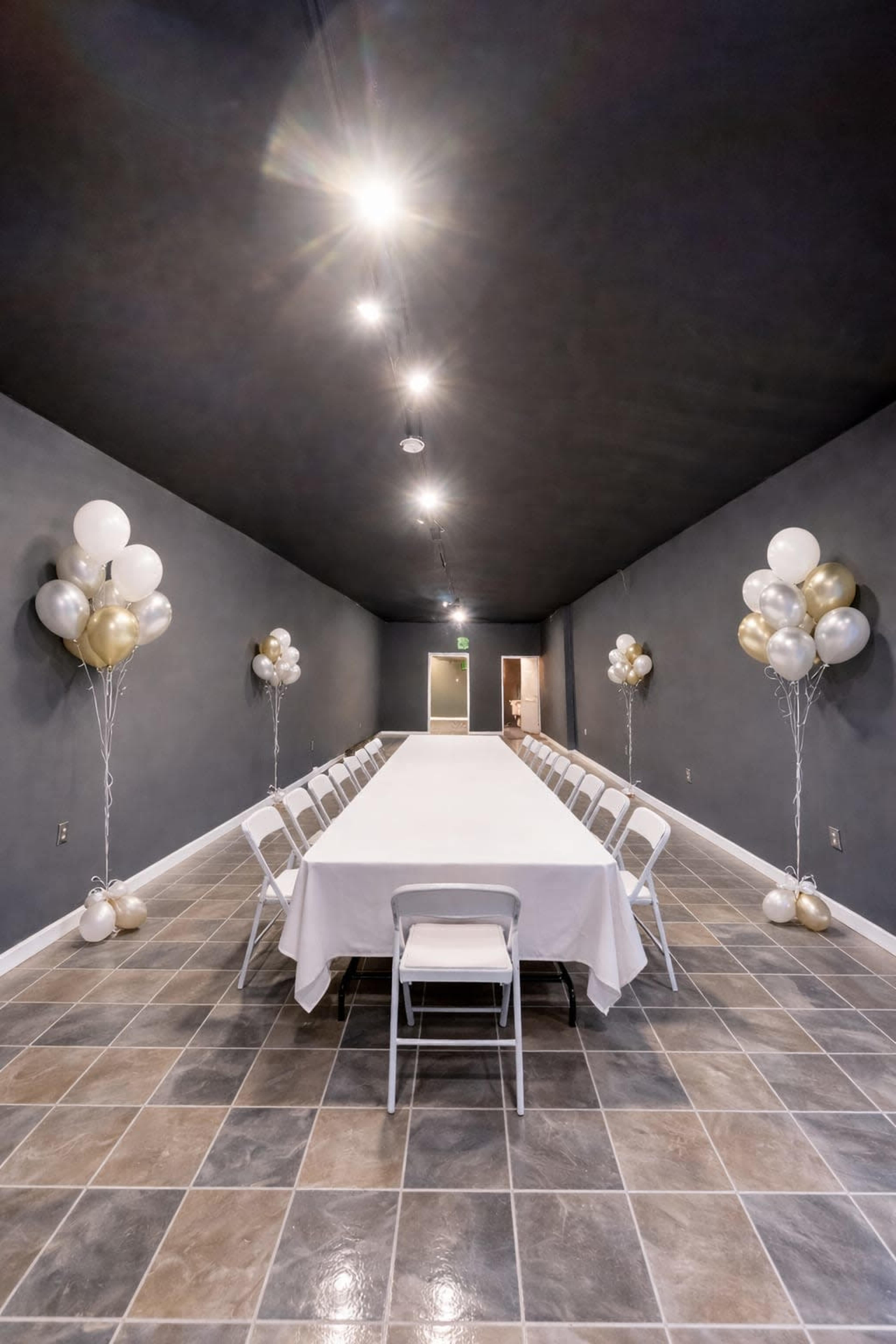 A long banquet table covered with a white tablecloth is set in a dimly lit room, flanked by balloon arrangements on either side.
