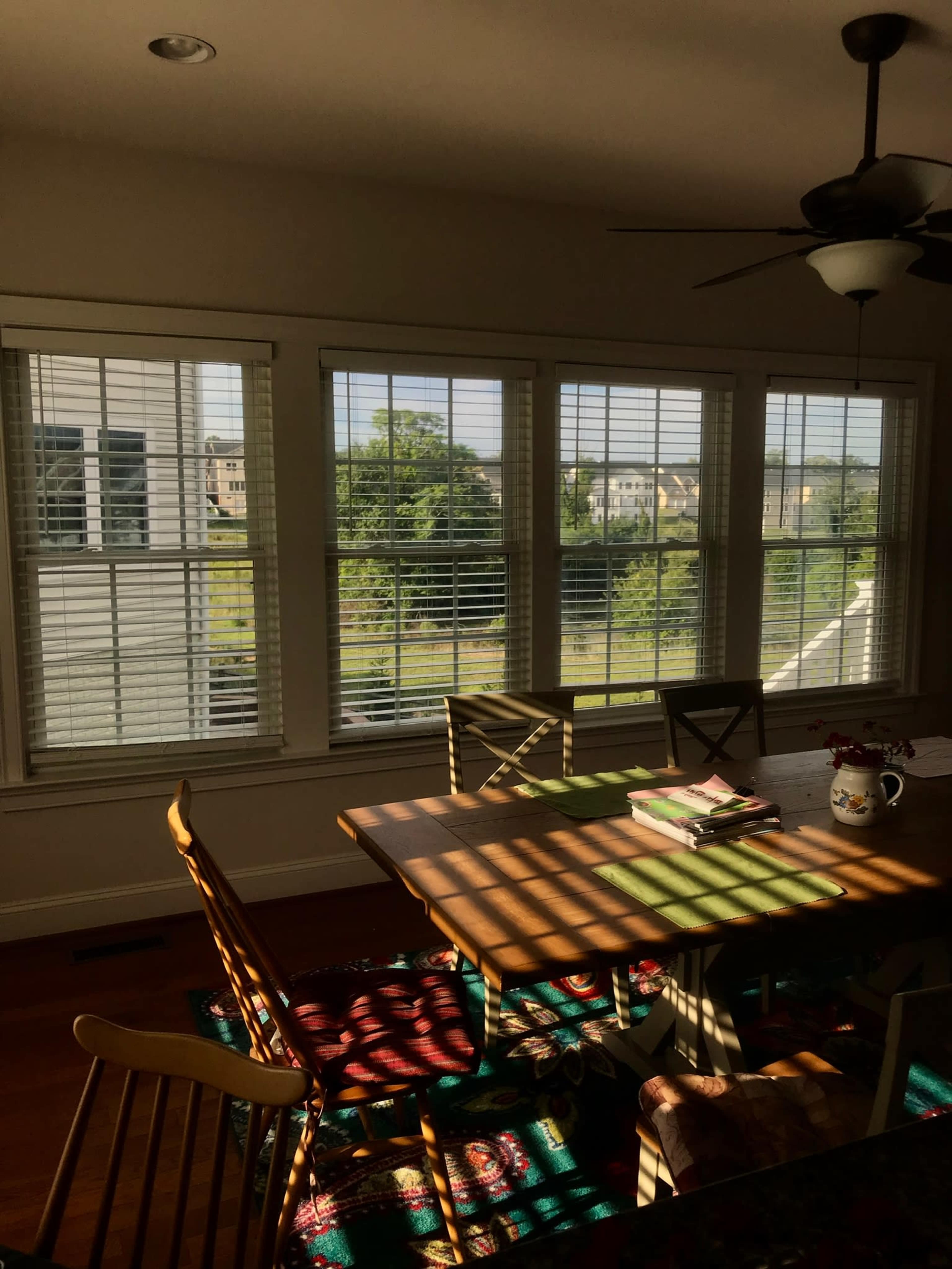 A dining area with a wooden table and chairs is illuminated by sunlight filtering through window blinds, casting shadows on the floor.