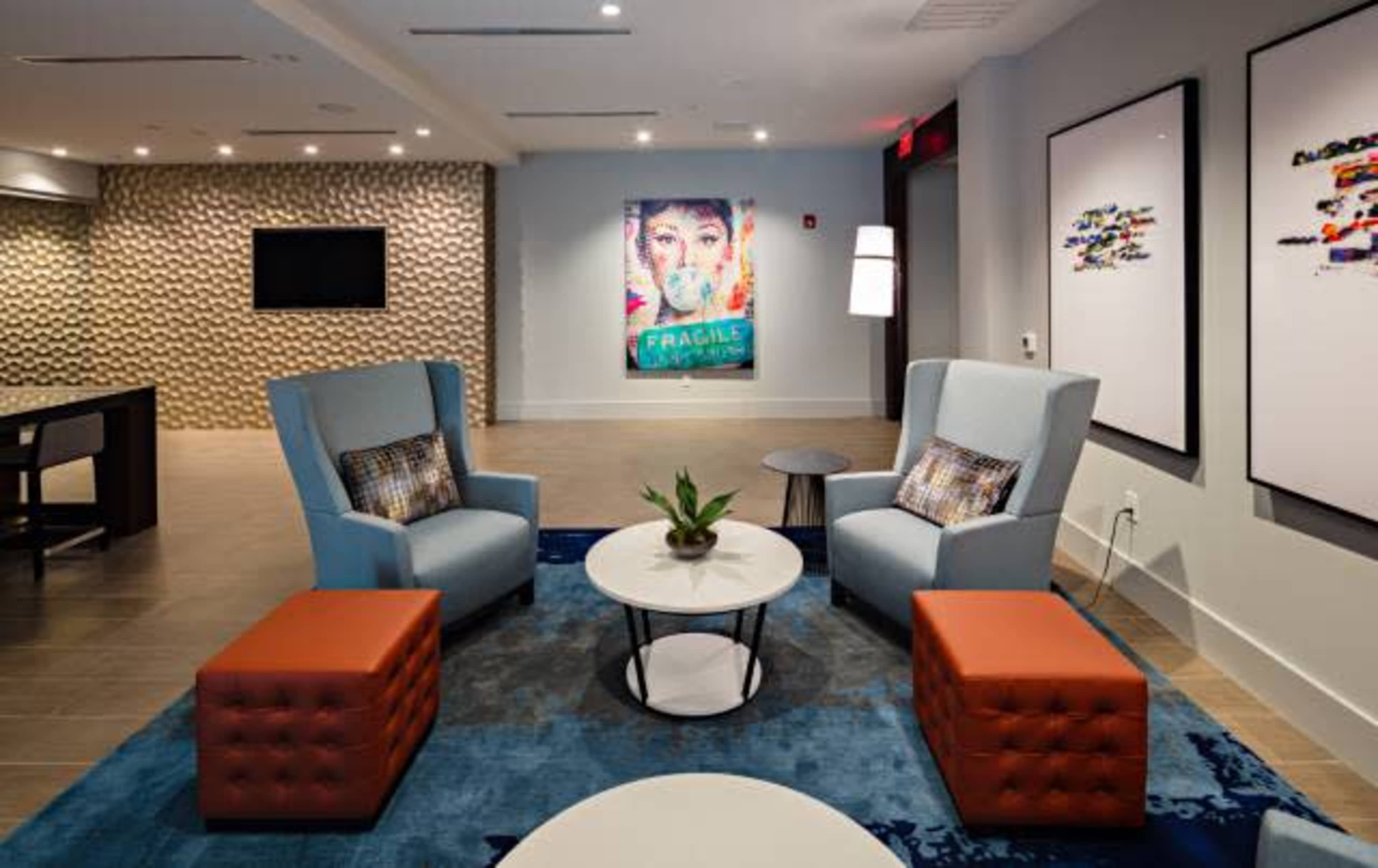 The image depicts a modern lounge area featuring two blue armchairs, a round table, and orange ottomans, with an artwork on the wall and textured design elements.