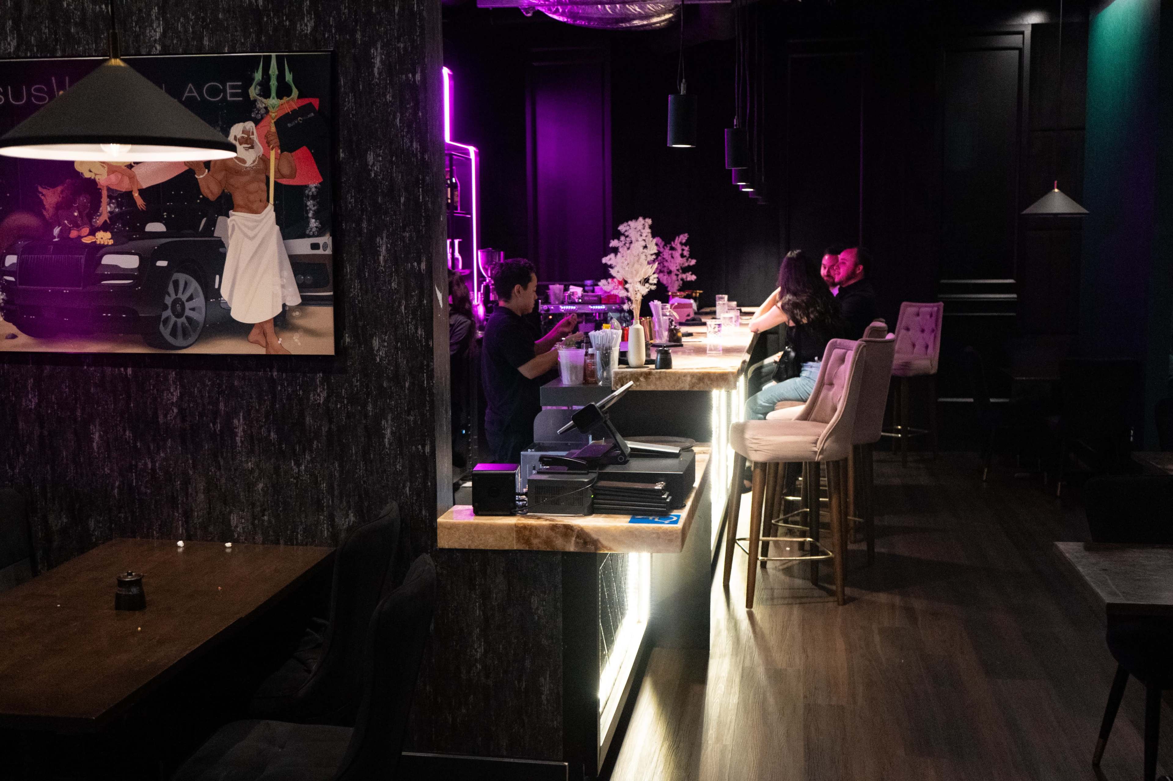 The image shows a modern bar interior with patrons sitting at the counter, illuminated by colorful lighting.
