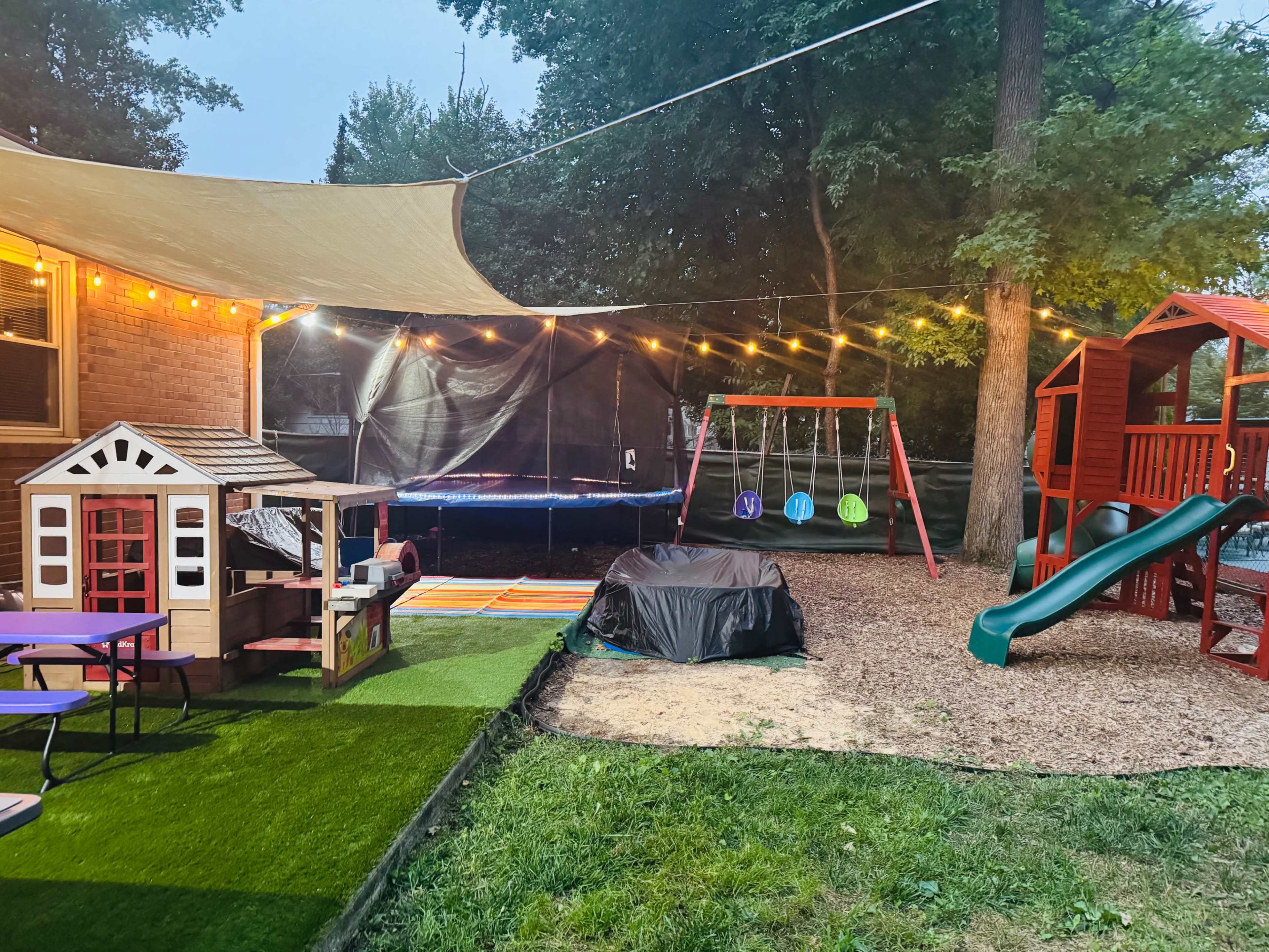 The image shows a backyard play area featuring a wooden playhouse, a trampoline, a swing set, a slide, decorative string lights, and a large shade sail overhead.