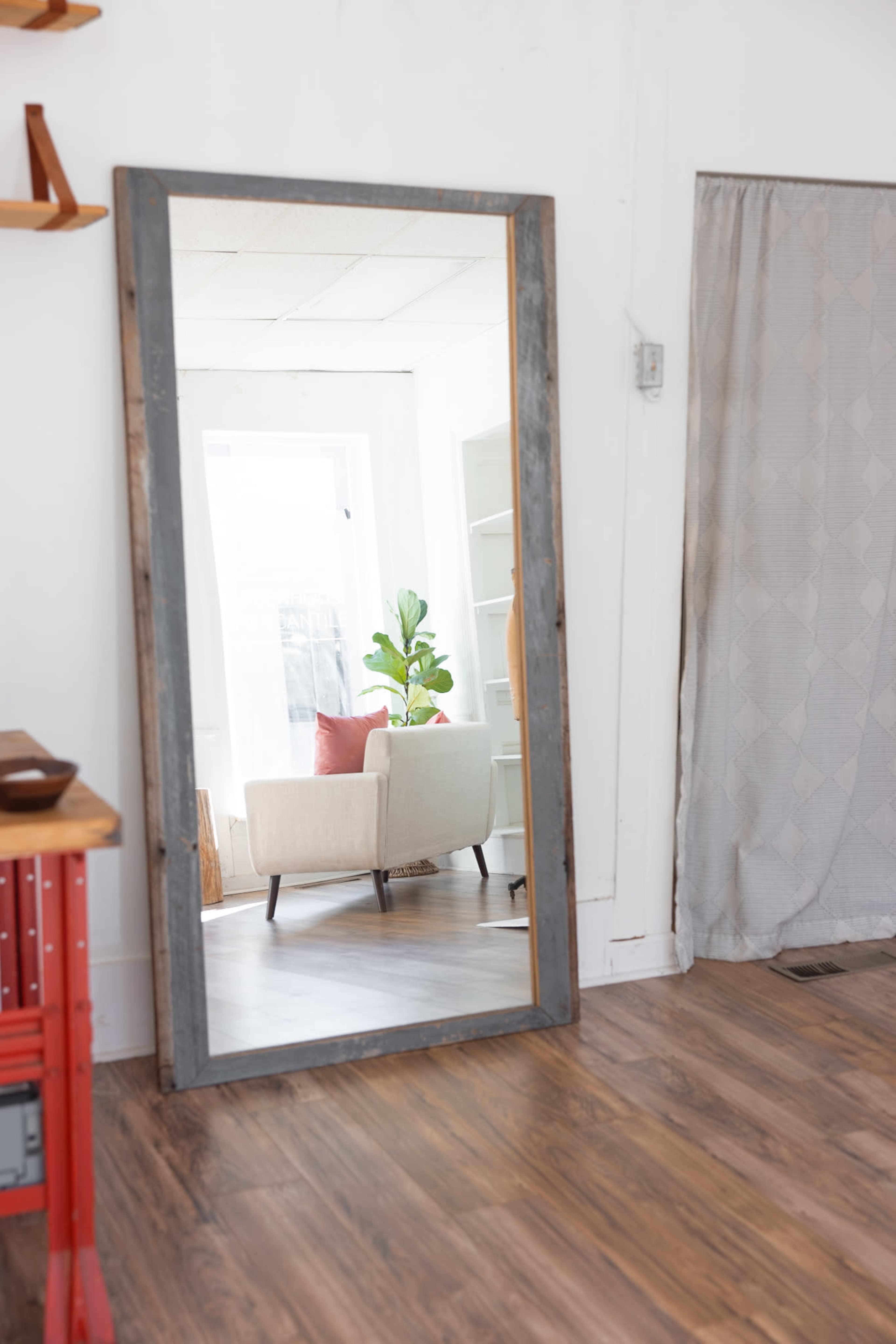 A large, rectangular mirror reflects a small, light-filled room with a white armchair and a potted plant.