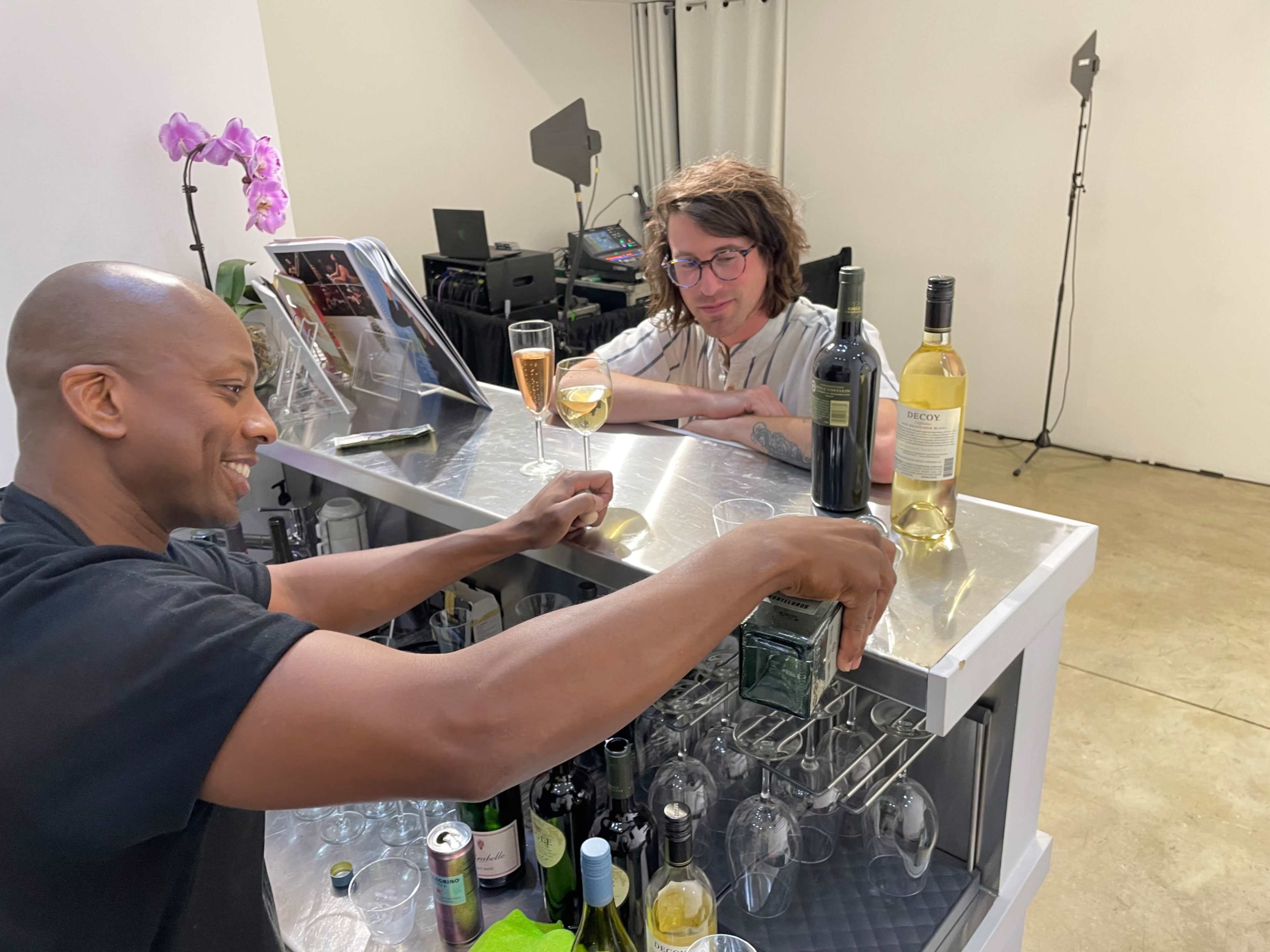 Two people are engaged at a bar counter, with one pouring a drink while the other observes, surrounded by various bottles and glasses.
