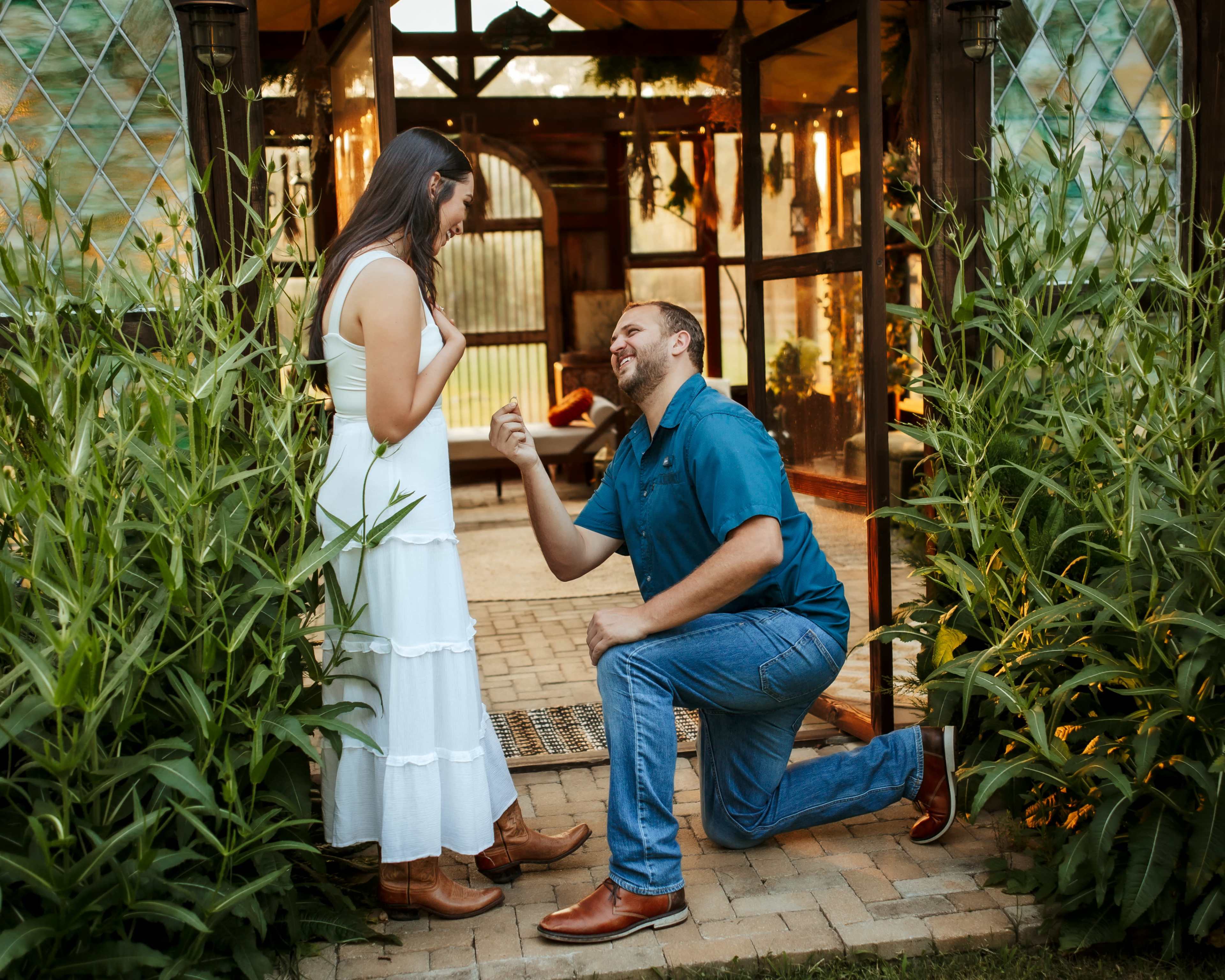 A man is kneeling in front of a woman in a white dress, proposing to her in a garden setting with greenery and a glasshouse in the background.