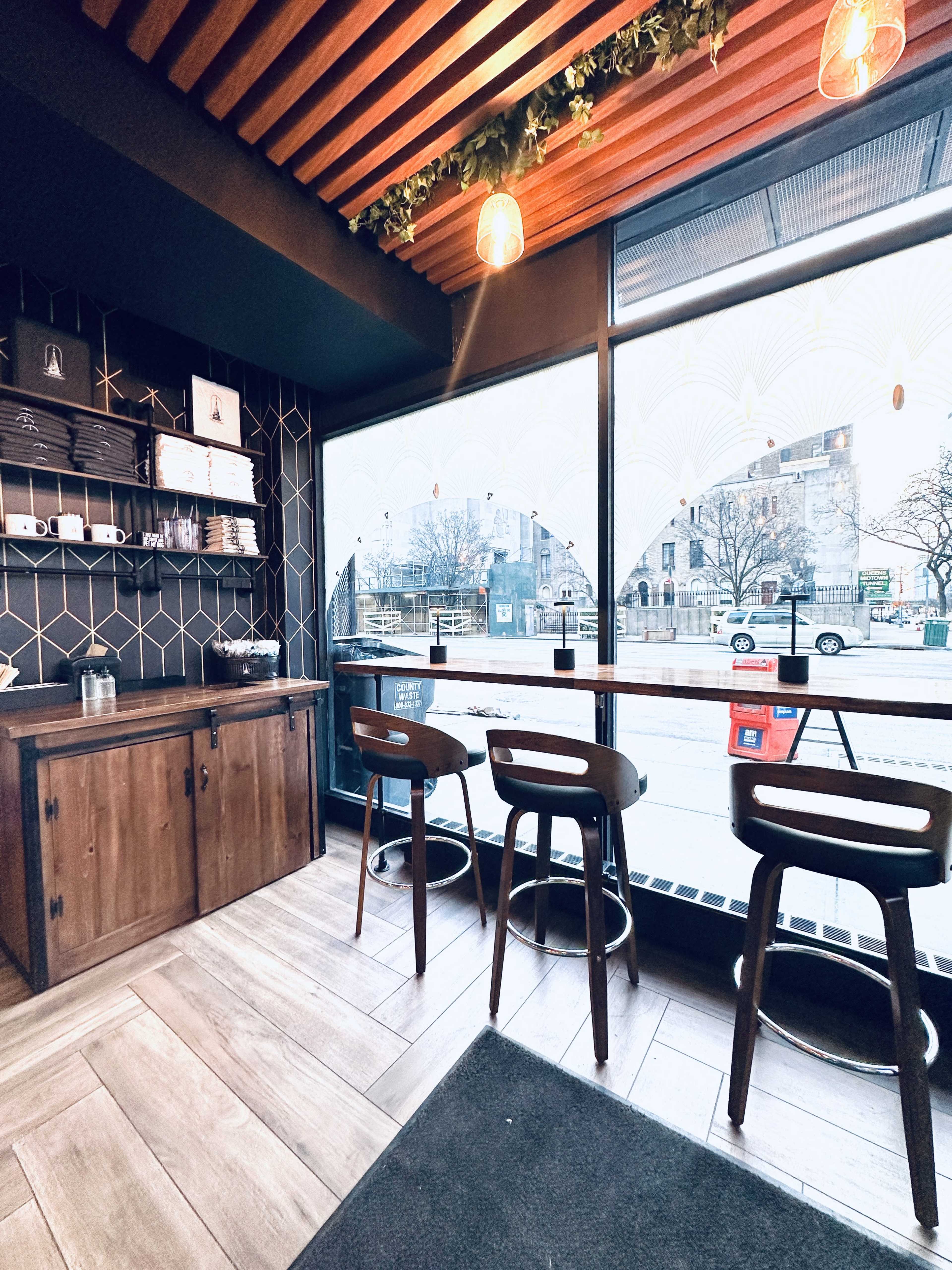 The image shows a cozy café interior featuring three wooden stools at a large window with decorative glass, and a wooden cabinet against the wall.