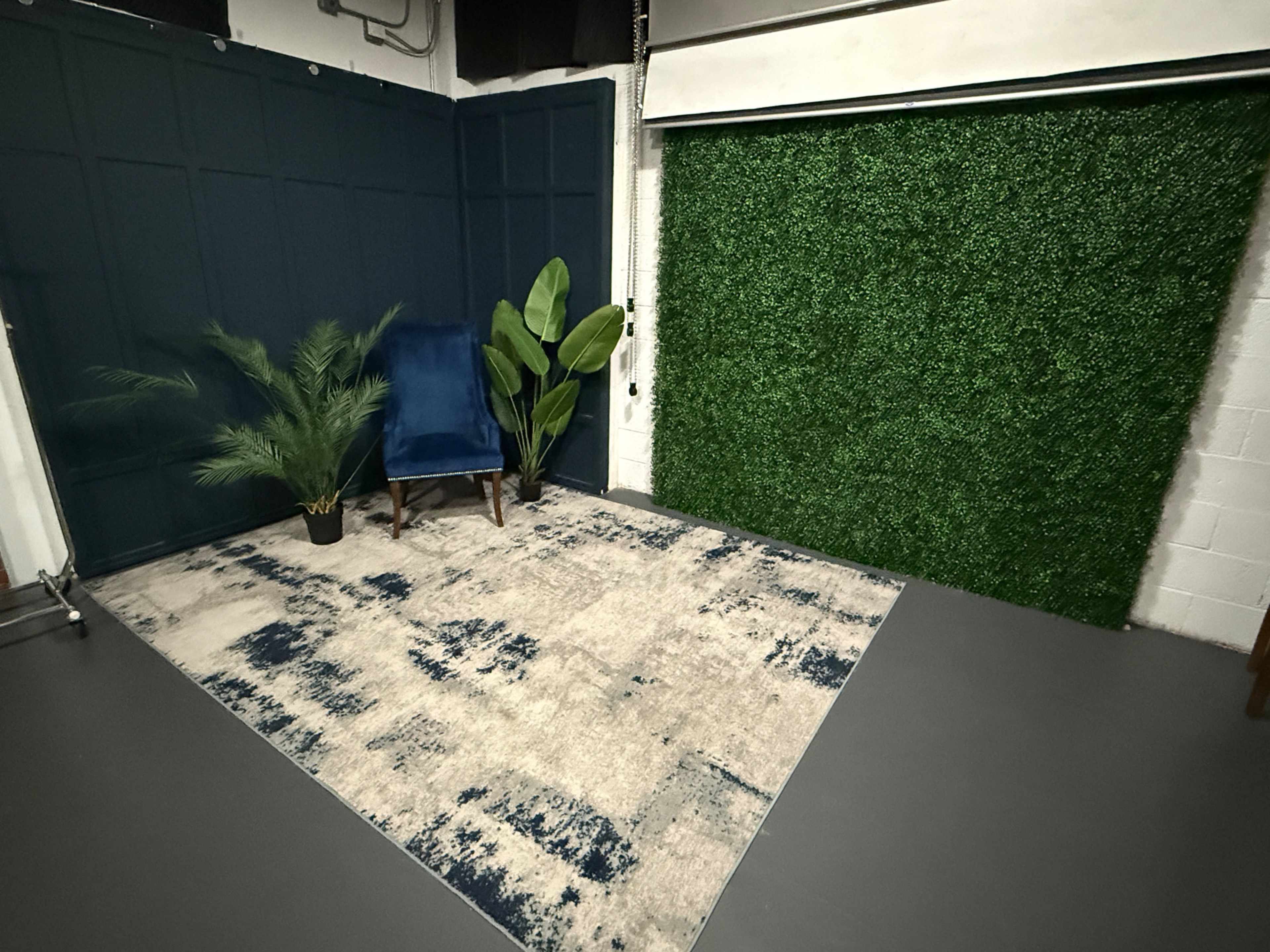 The image shows a room with a green wall of artificial foliage, a blue armchair, a potted plant, and an area rug on a gray floor.