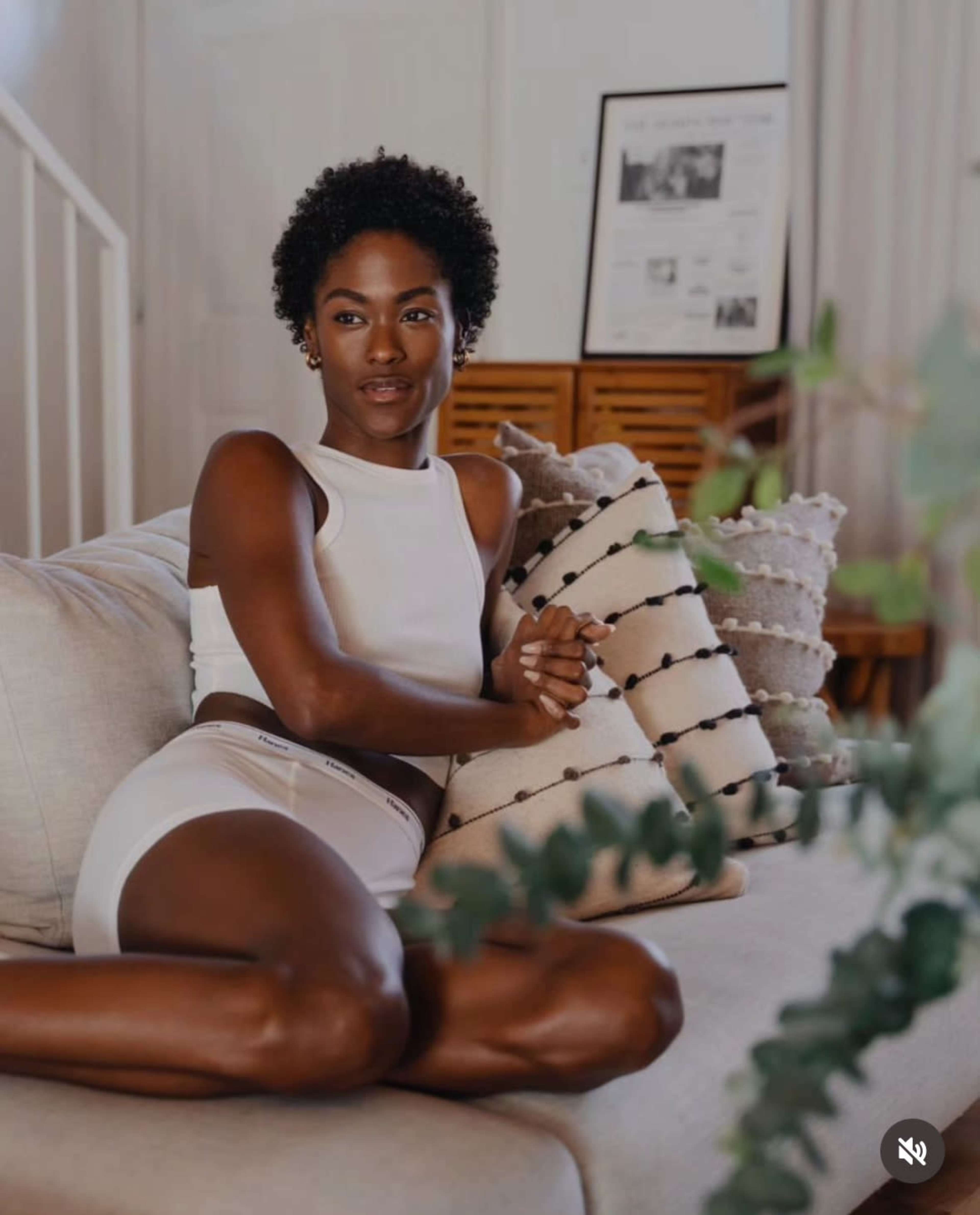 A person with short curly hair sits on a beige sofa, wearing a white crop top and white shorts, with decorative pillows beside them.
