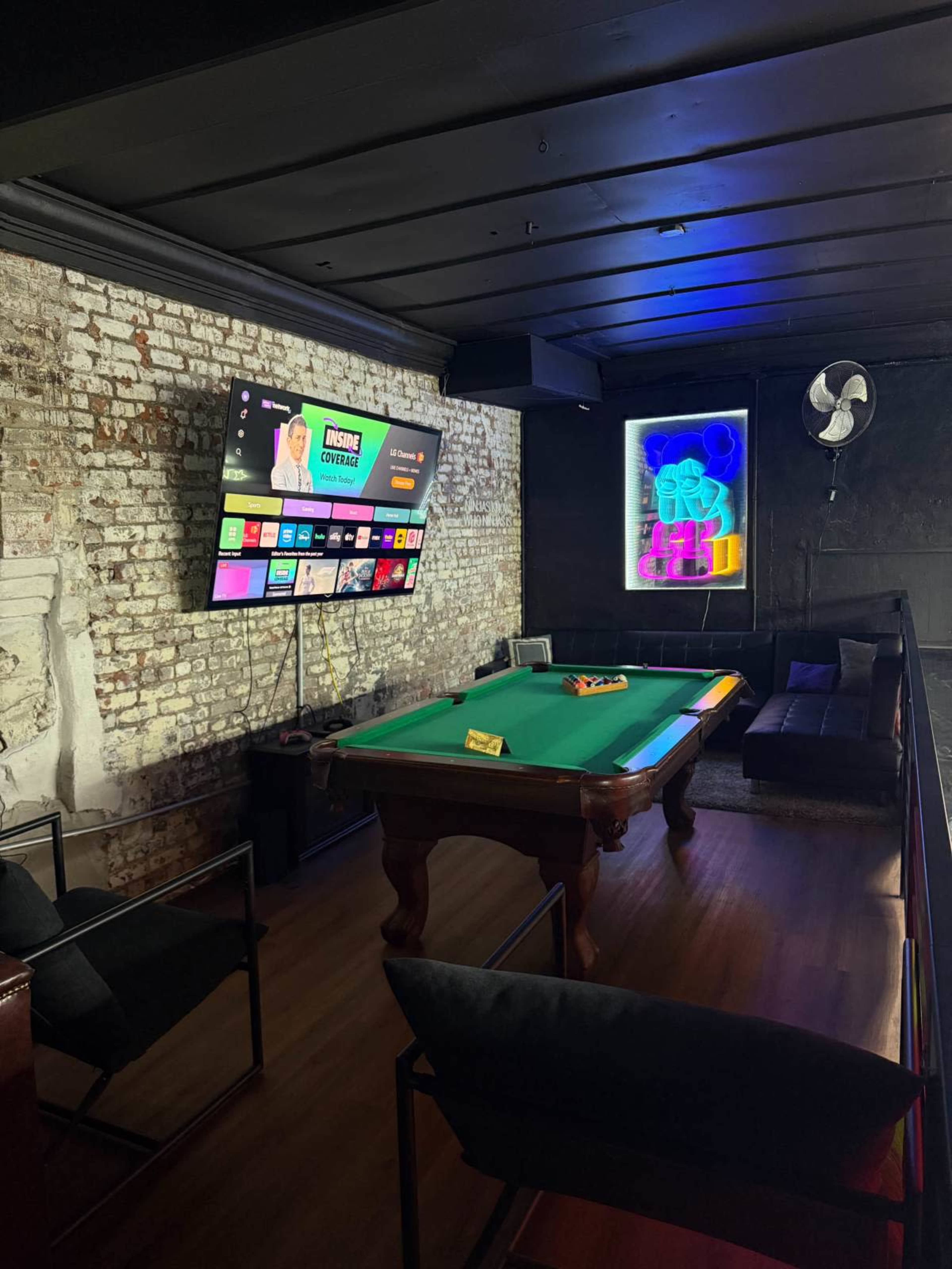 The image shows a game room featuring a billiards table, a large television mounted on a brick wall, and a cozy seating area illuminated by neon lights.