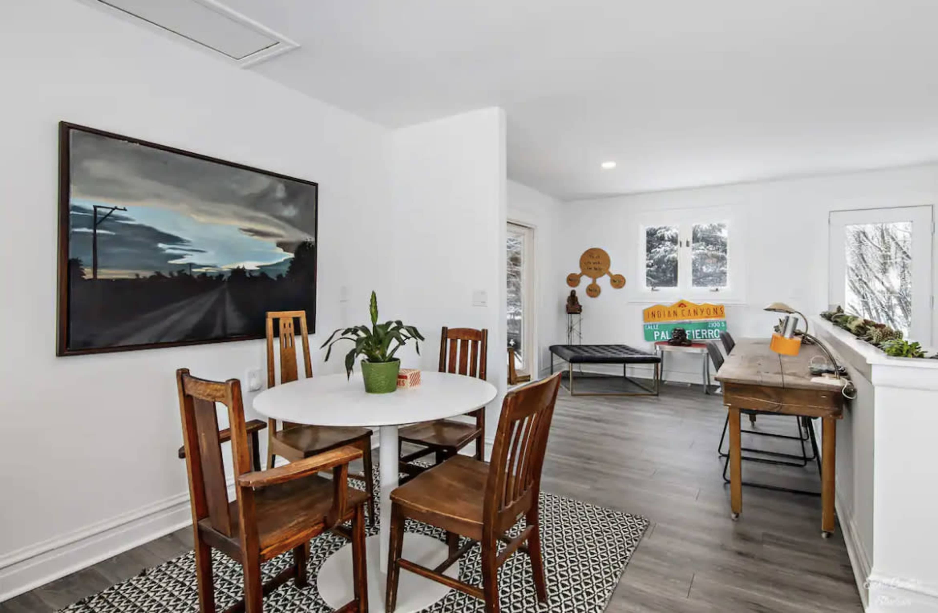 A dining area features a round table surrounded by four wooden chairs, with a dark landscape painting on the wall and a bright, open space in the background.